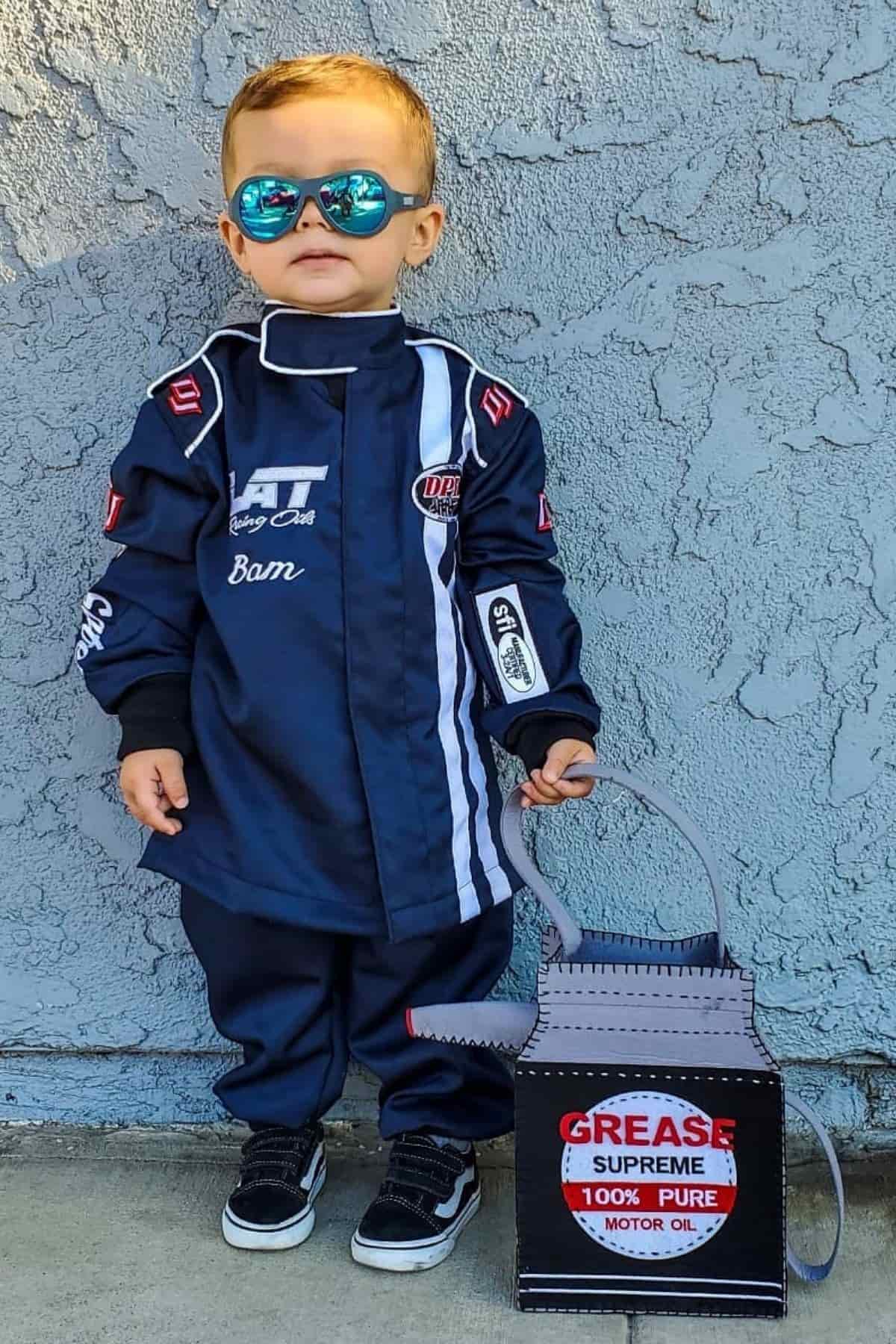 Toddler in a racing suit and sunglasses stands next to a box labeled “Grease Supreme 100% Pure Motor Oil” against a textured wall.