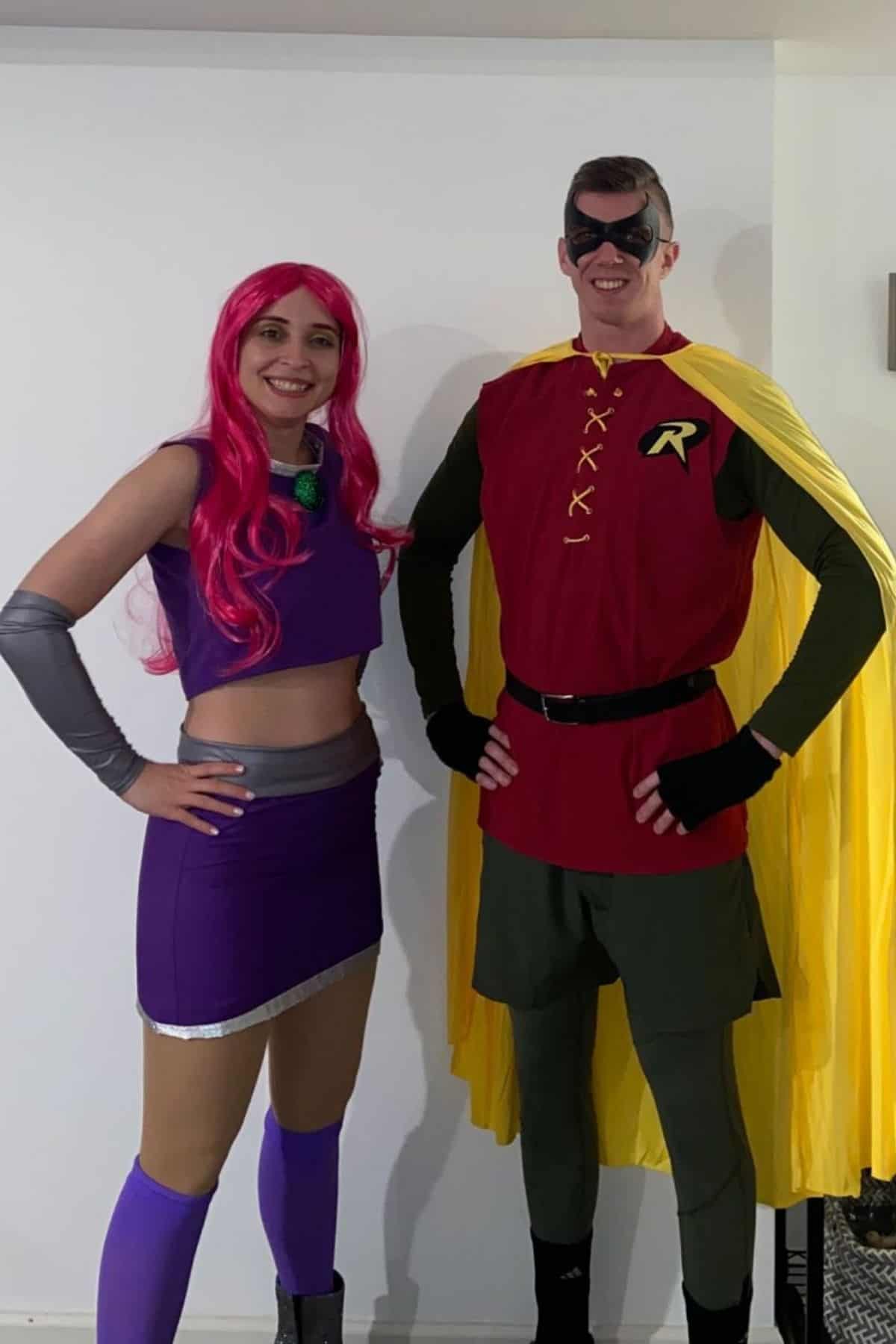 Two people pose in superhero costumes; one as Starfire in a purple outfit and pink wig, and the other as Robin in a red, green, and yellow costume with a cape and mask, standing indoors.