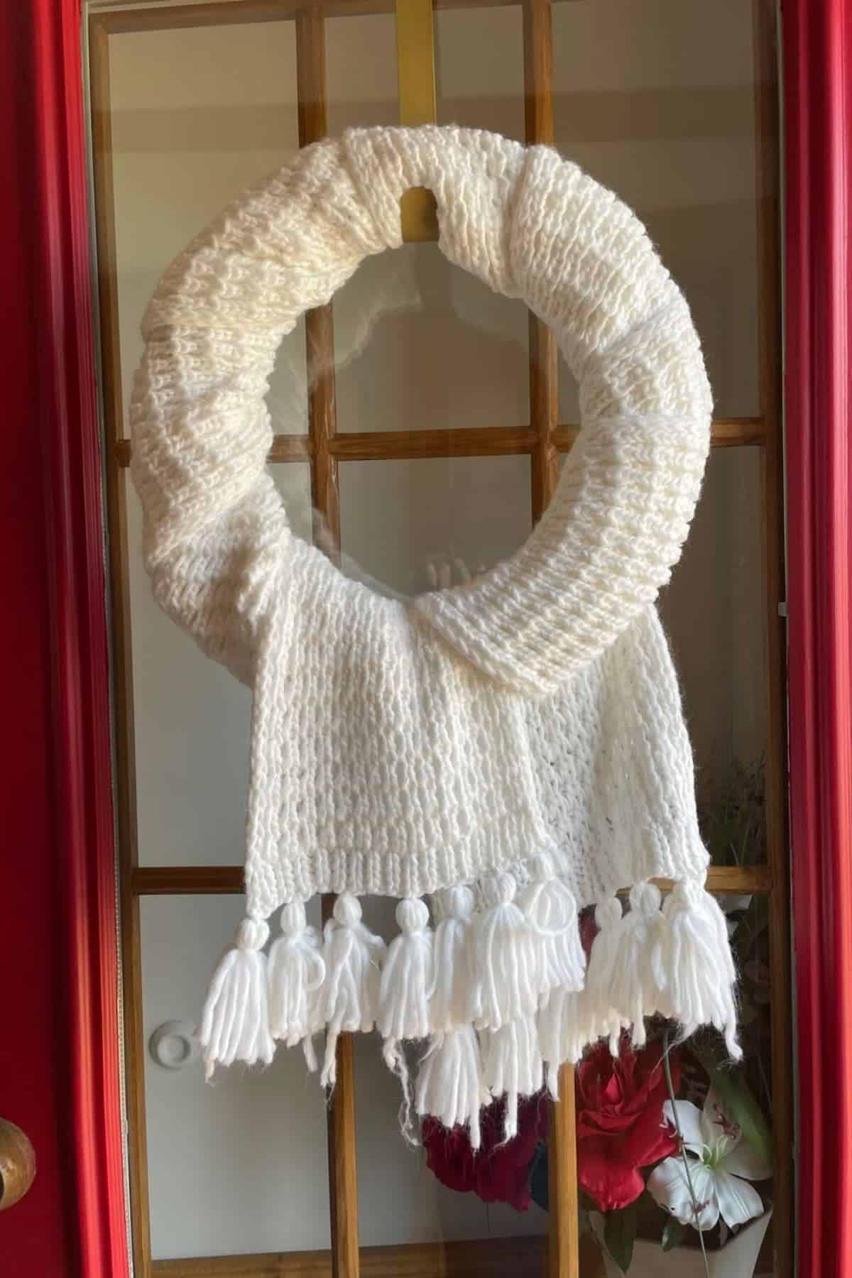 A white knitted scarf is wrapped around a round wreath form and hangs on a glass door, with tassels hanging at the bottom.