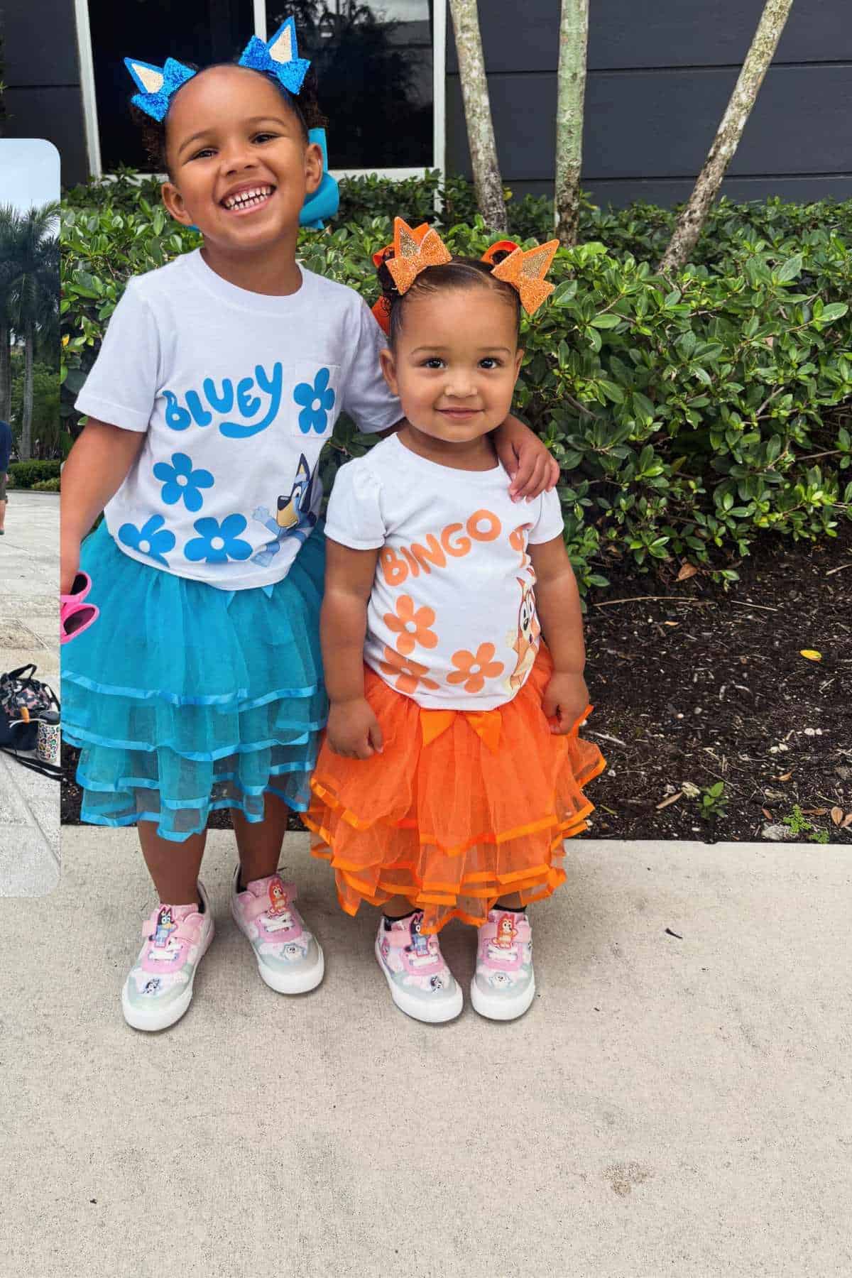 Two young children stand outside, smiling. One wears a Bluey shirt and blue skirt, and the other wears a Bingo shirt and orange skirt. Both have matching headbands and sneakers.