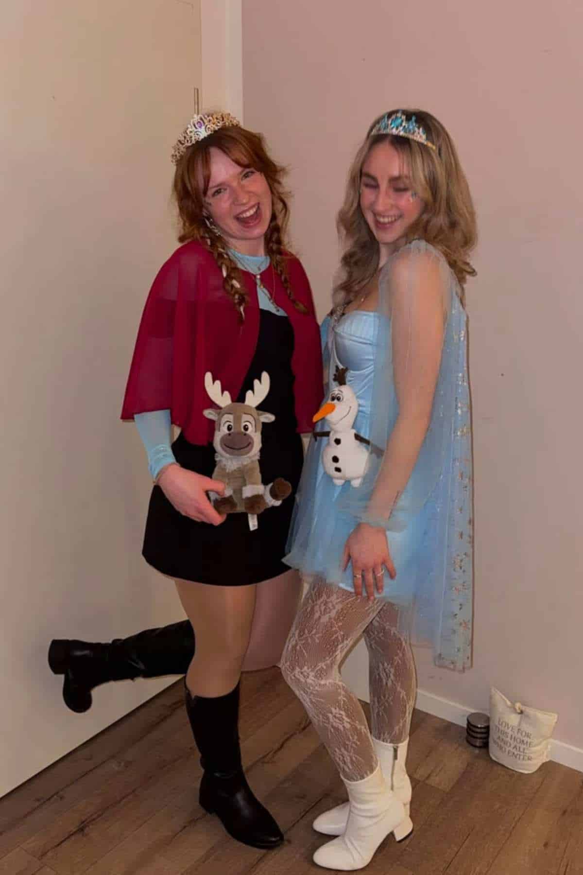 Two people pose indoors wearing Frozen-inspired costumes, one dressed as Anna holding a Sven plush, the other as Elsa holding an Olaf plush. Both are smiling and wearing tiaras.