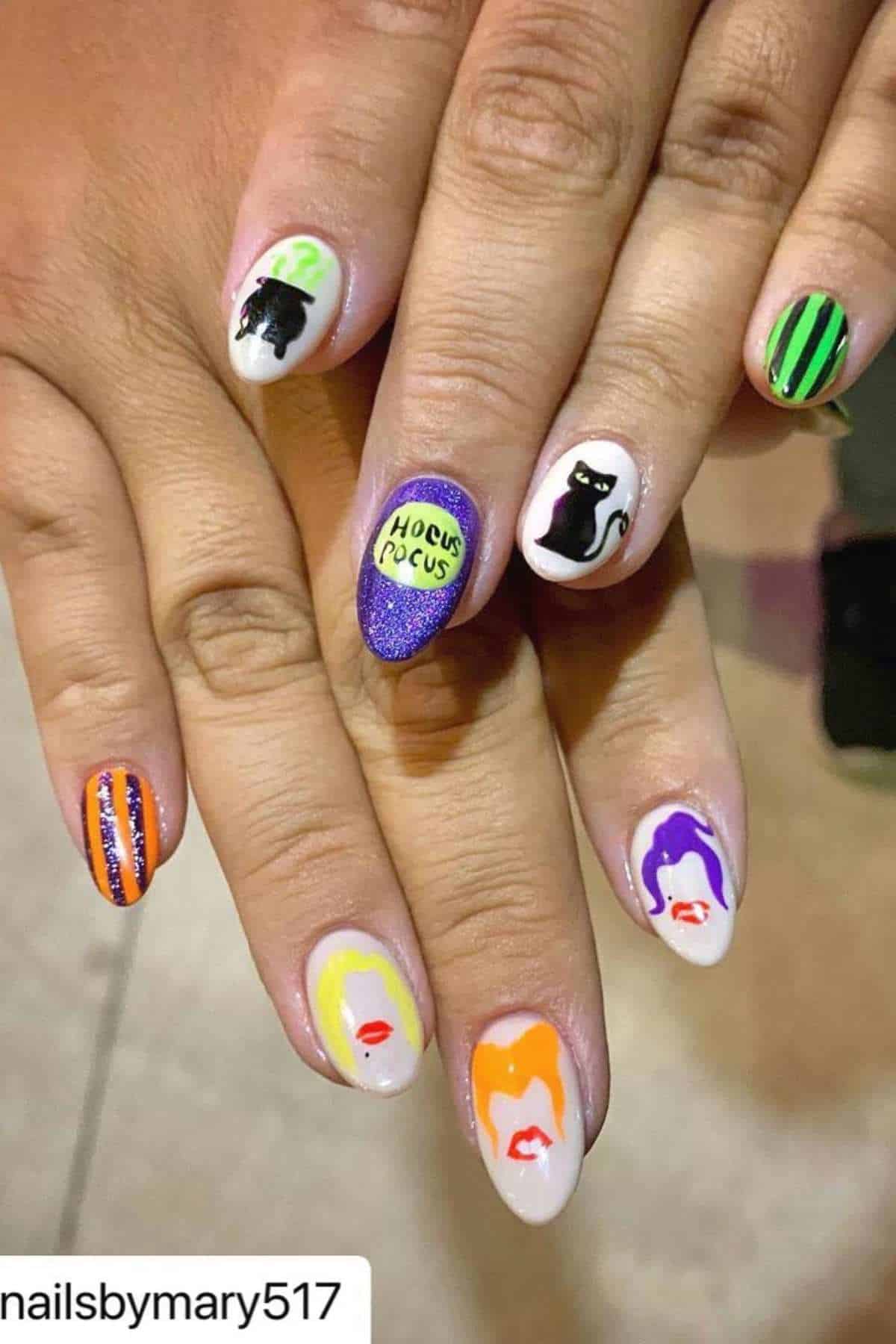 Hands with Halloween-themed nail art featuring witches’ faces, a black cat, the words “Hocus Pocus,” and striped patterns in black, green, orange, and purple on a neutral base.