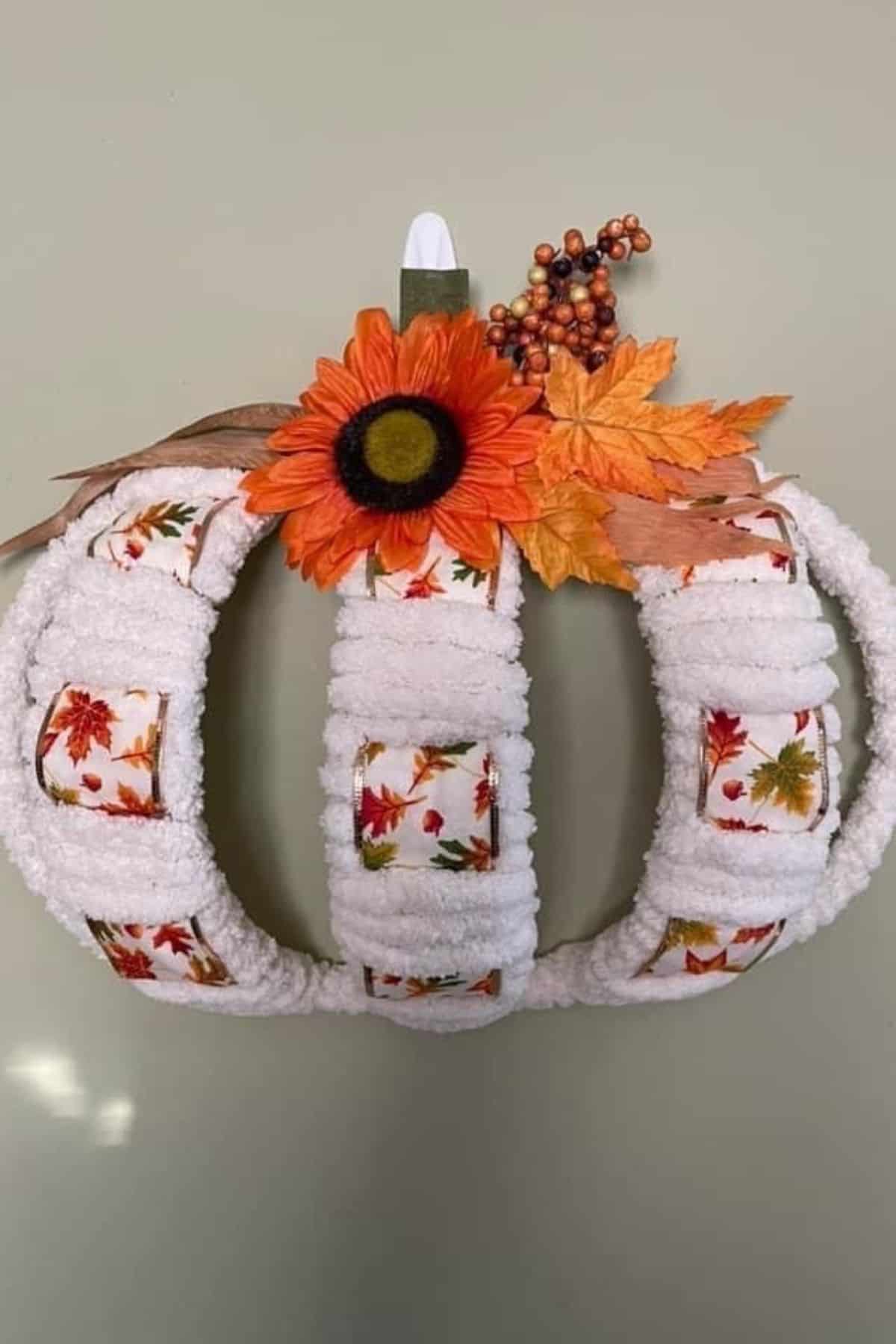 A decorative pumpkin-shaped wreath made of white fabric and autumn leaf-patterned ribbon, adorned with a faux orange sunflower and fall leaves.