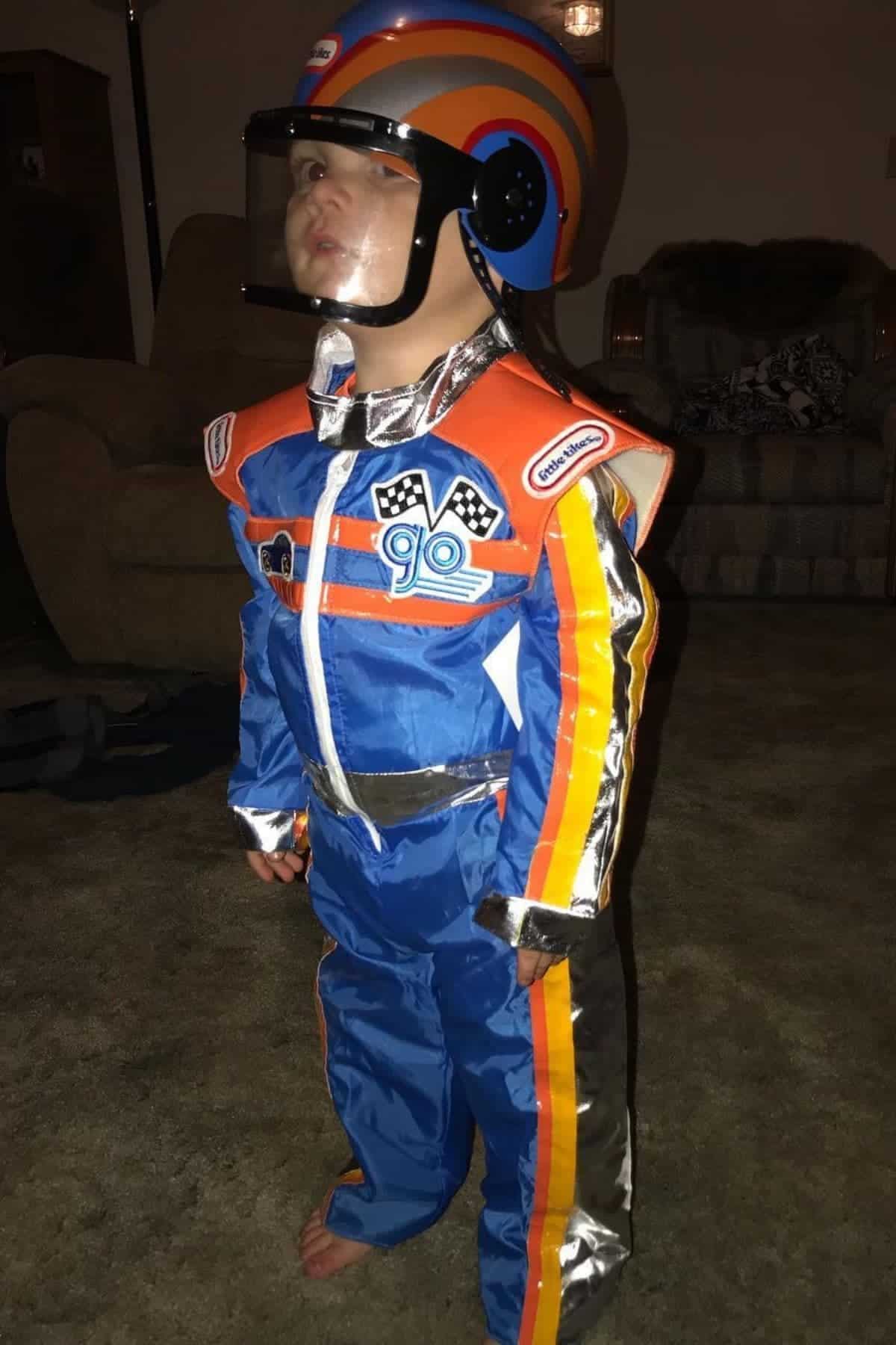 A young child in a blue racing jumpsuit and helmet stands indoors on a carpeted floor, looking upward with a serious expression.
