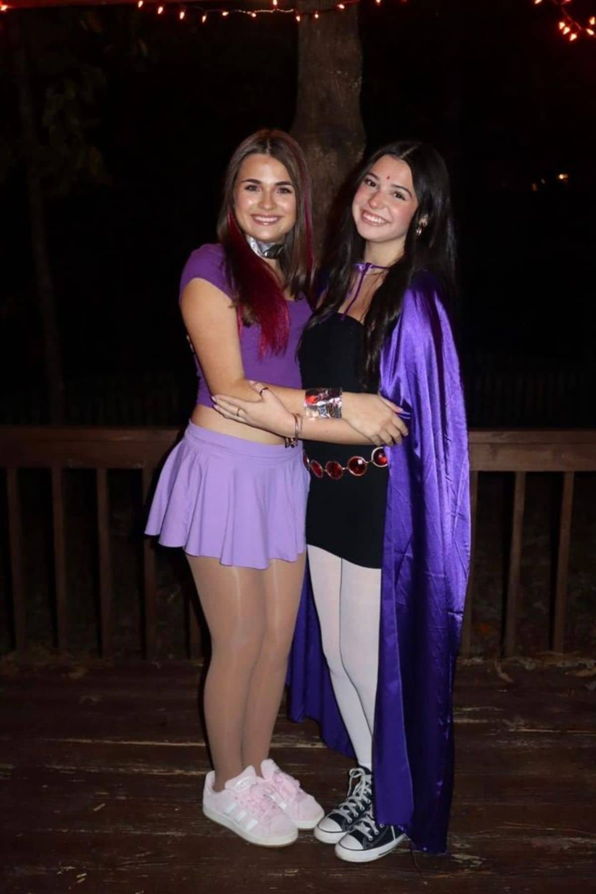 Two young women in purple costumes pose together outside at night, standing on a wooden deck with string lights above them.