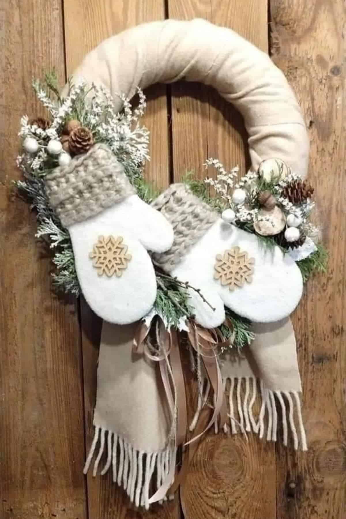 A wreath decorated with white mittens, greenery, pinecones, white berries, and a beige scarf hangs on a wooden door.