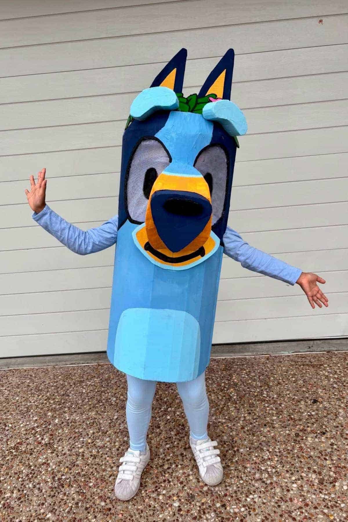 Person wearing a large handmade Bluey costume, standing on a pebble path with arms outstretched in front of a white garage door.