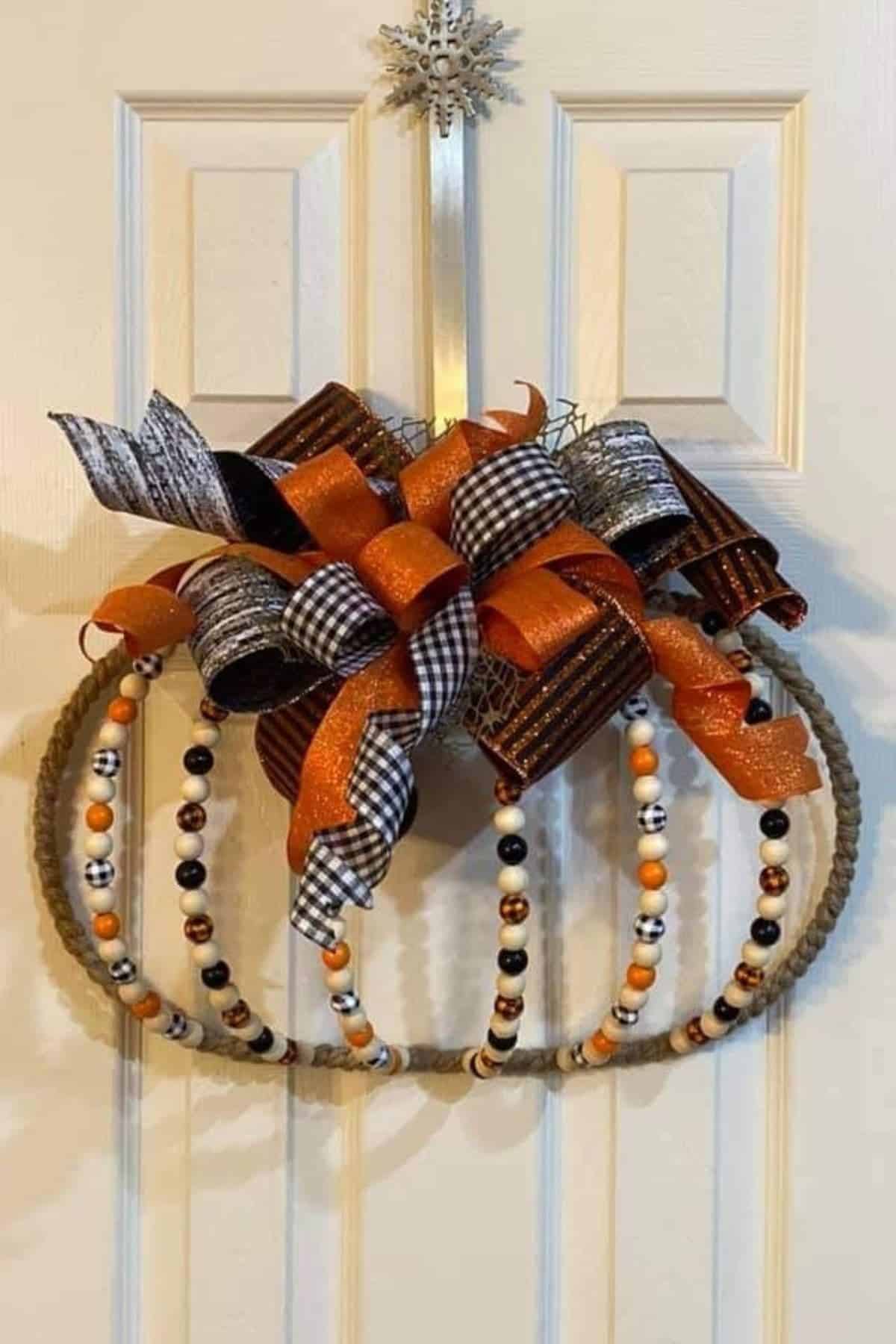 A pumpkin-shaped door wreath made of rope and beaded wire, decorated with orange, black, and white bows, hangs on a white paneled door.