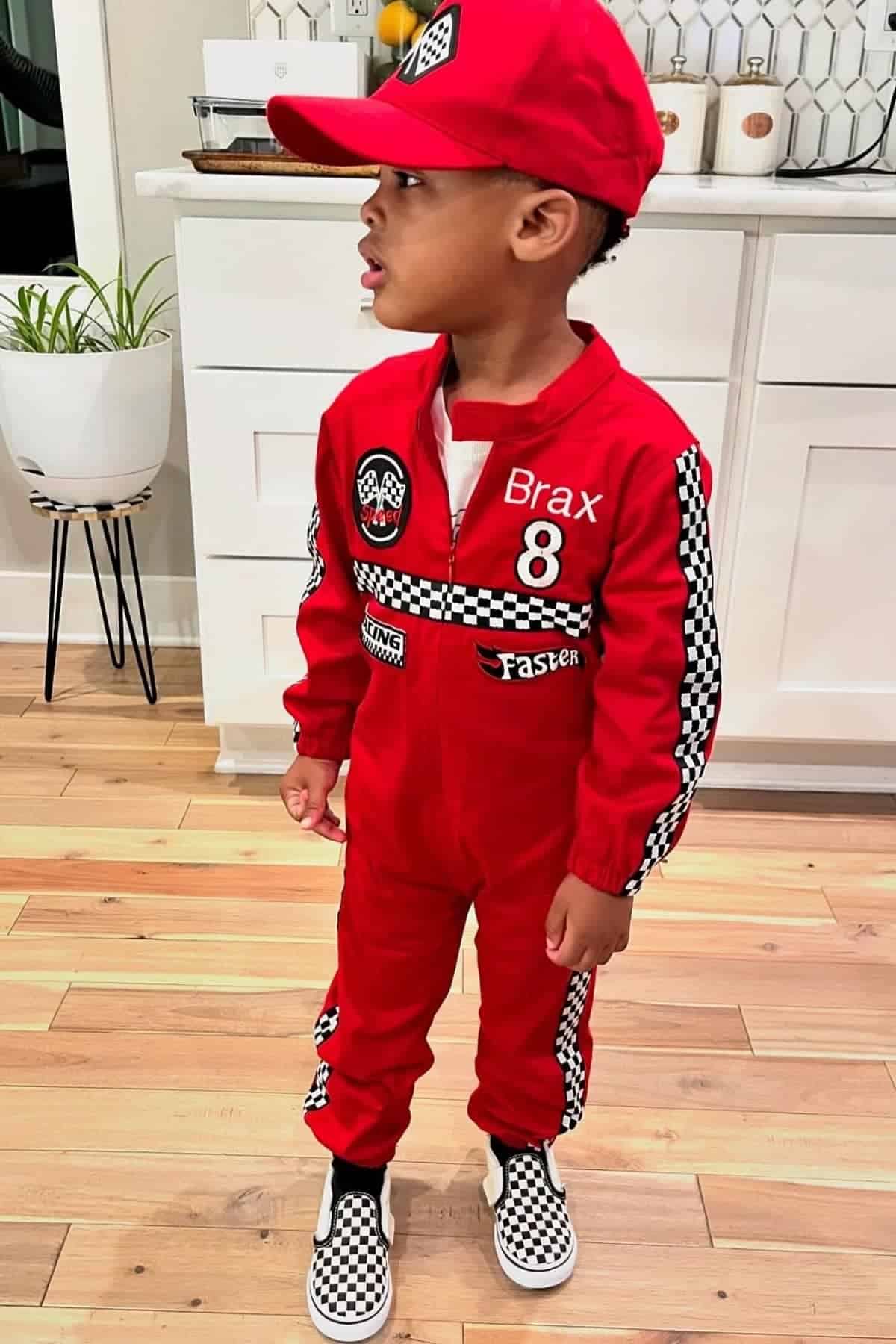 A young child in a red racing suit and matching cap stands indoors on a wooden floor, looking to the side. The suit features checkered patterns and racing-themed patches.