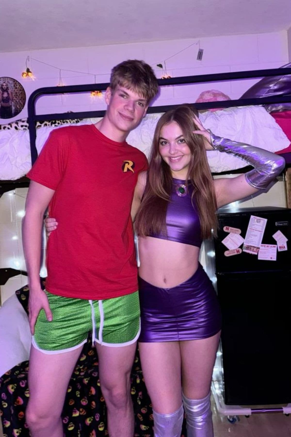 Two people pose in front of a bunk bed. One wears a red “R” t-shirt and green shorts; the other wears a shiny purple crop top and skirt with silver accessories.