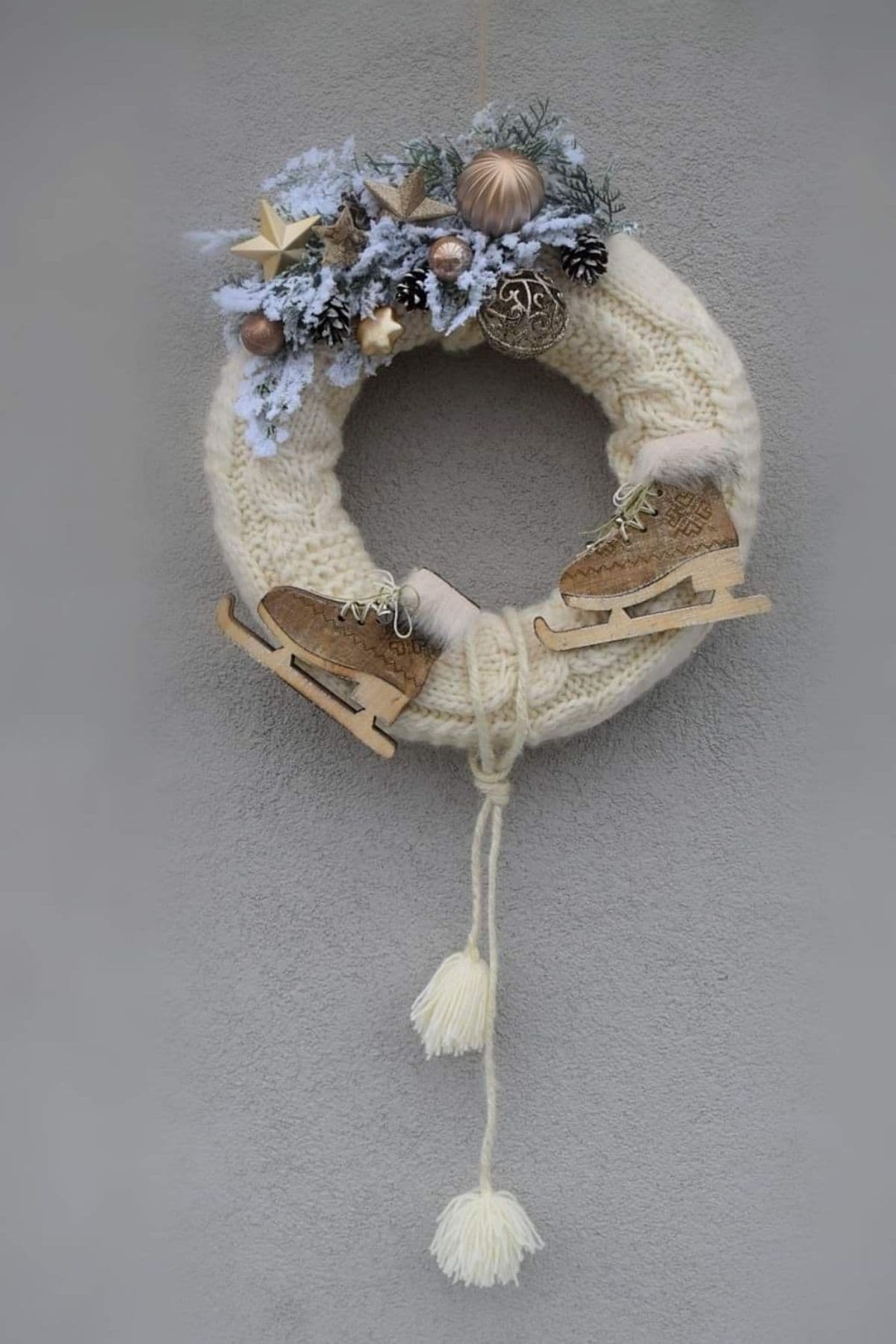A cream-colored knit wreath decorated with pinecones, baubles, faux greenery, wooden ice skates, and two yarn tassels, hanging on a gray wall.