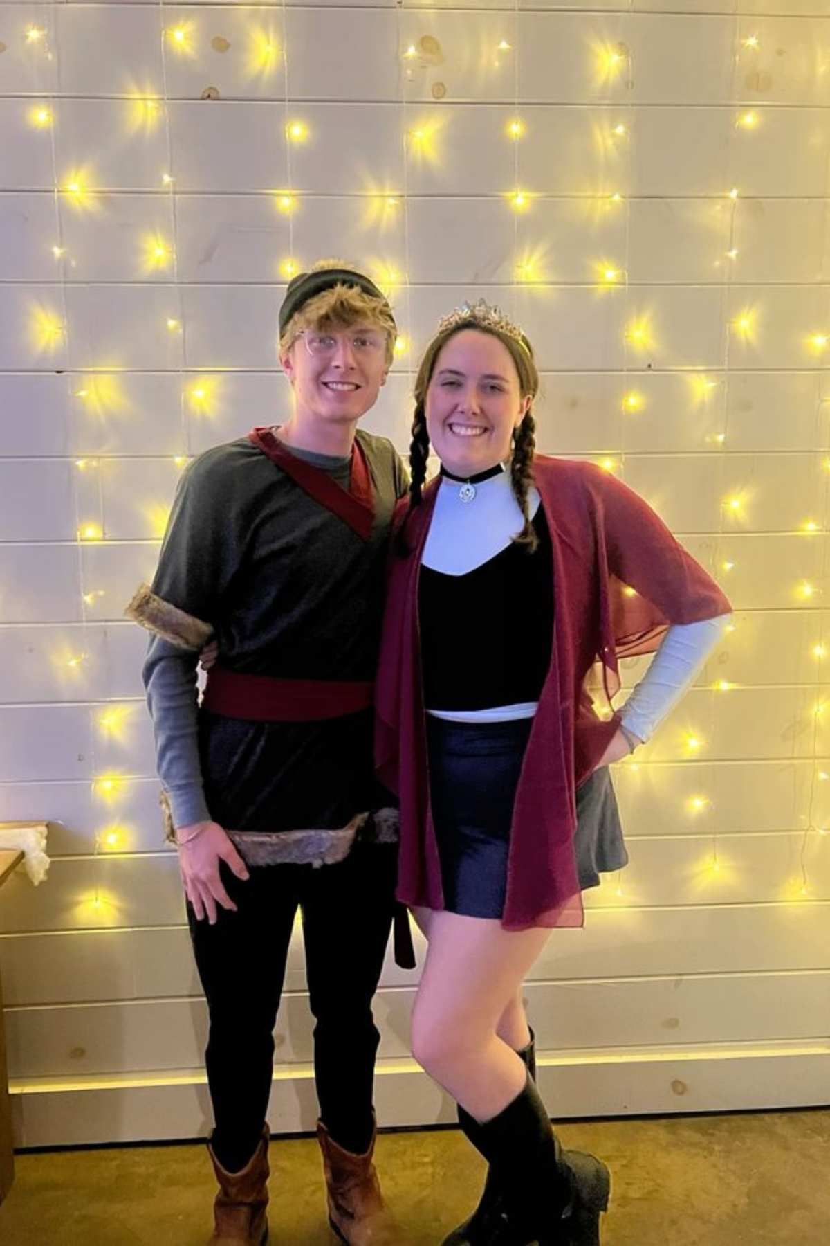 Two people in costumes stand side by side, smiling in front of a white wooden wall decorated with warm string lights.