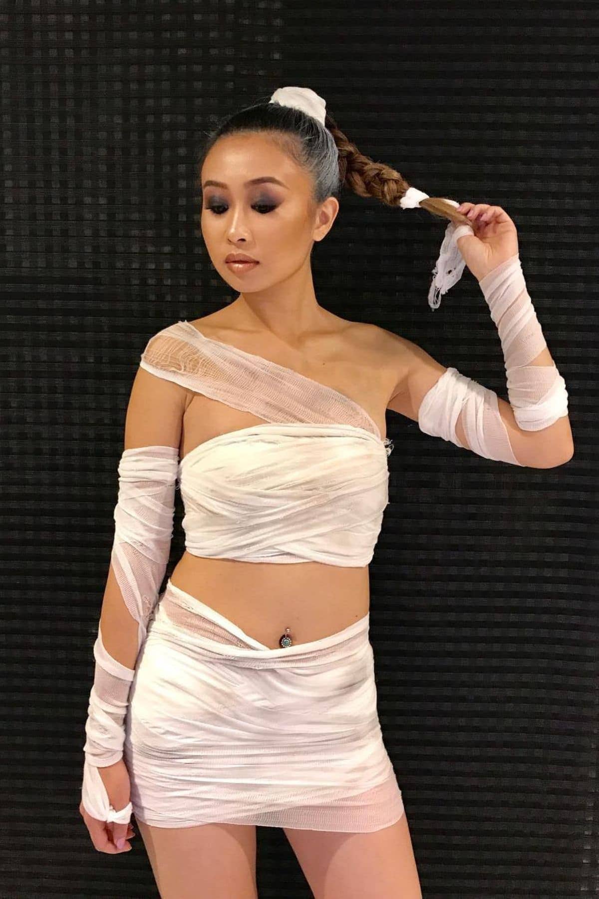 Woman wearing a white, one-shoulder, wrap-style outfit with sheer fabric, holding her braided ponytail, standing against a black textured background.