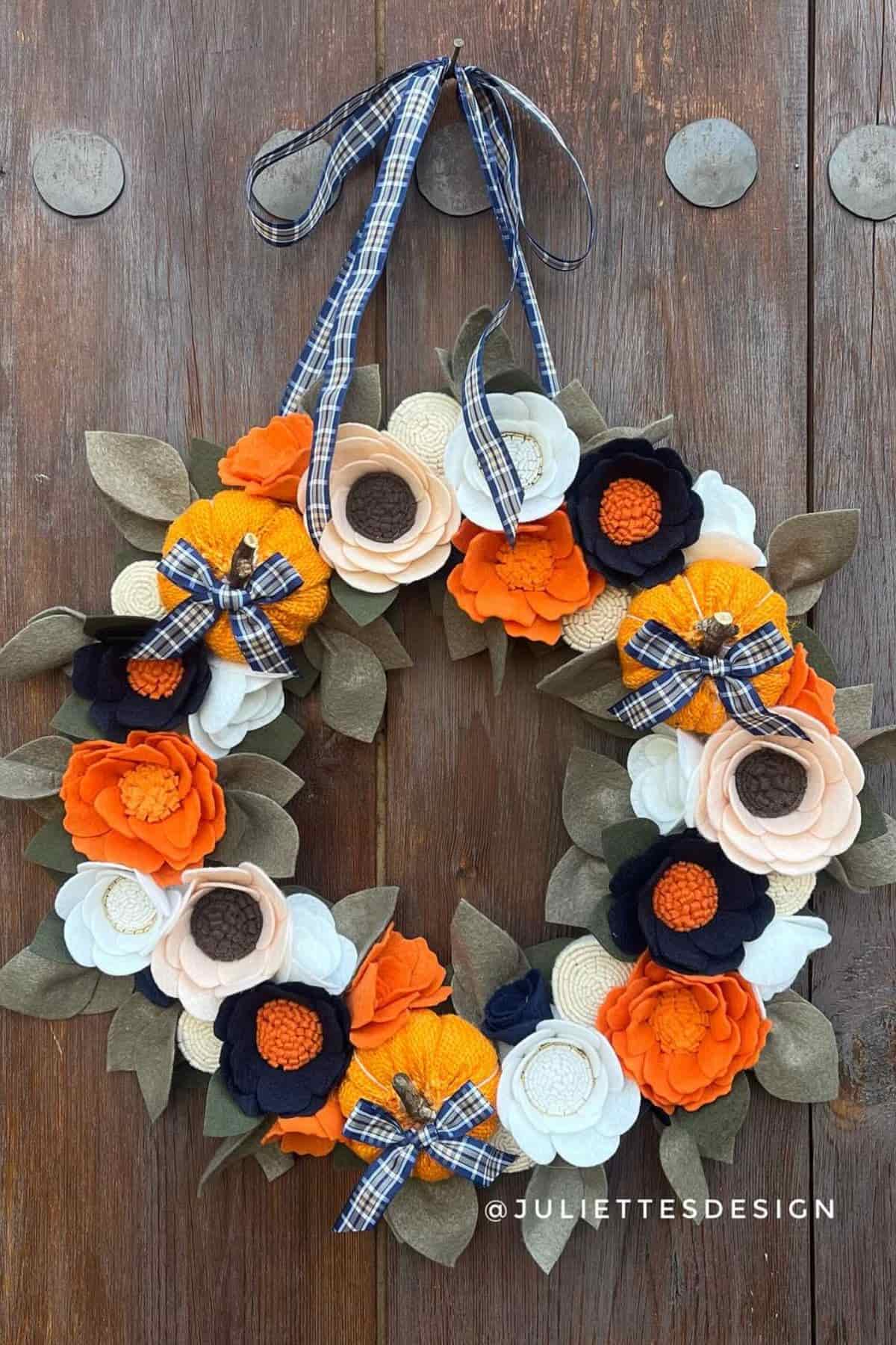 A decorative wreath with orange, cream, black, and beige fabric flowers, plaid bows, and green felt leaves, hanging on a wooden door.