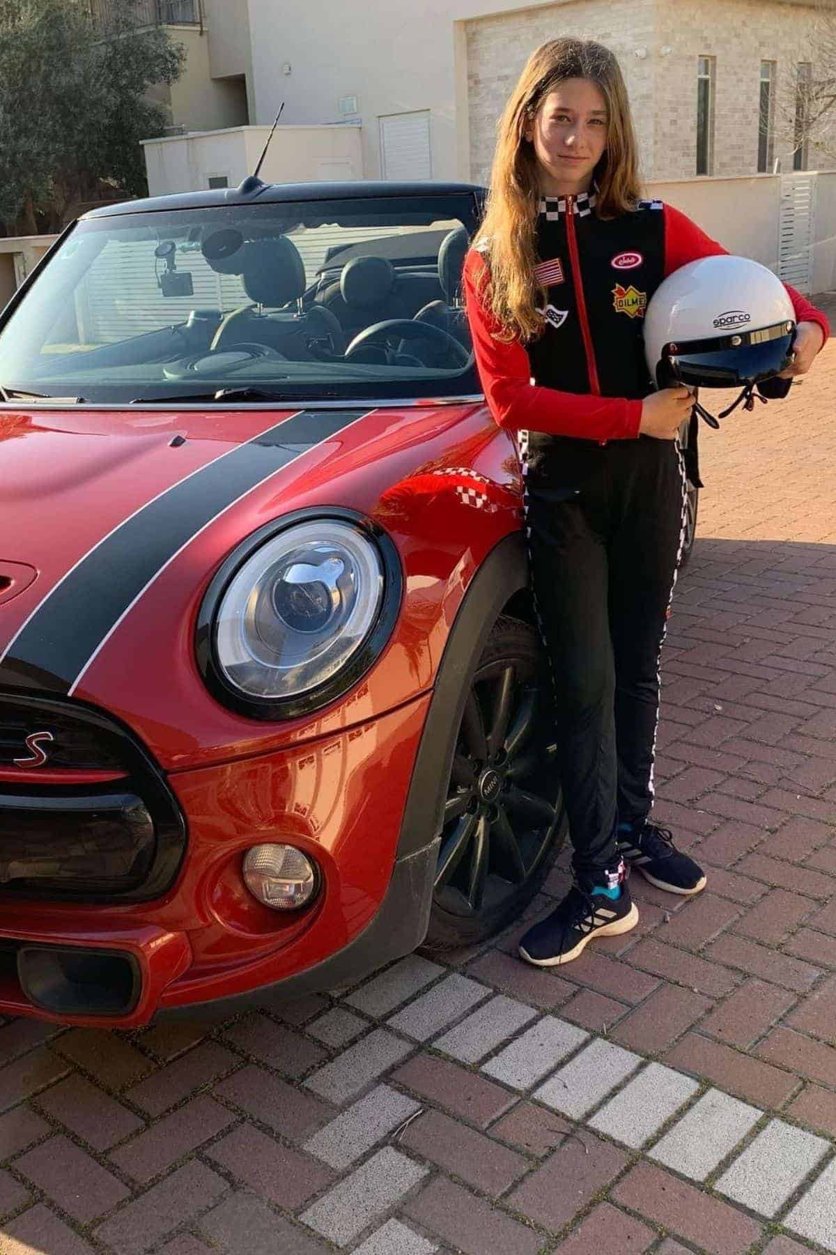 A young person in a racing suit holds a helmet and stands next to a red Mini Cooper convertible parked on a brick driveway.