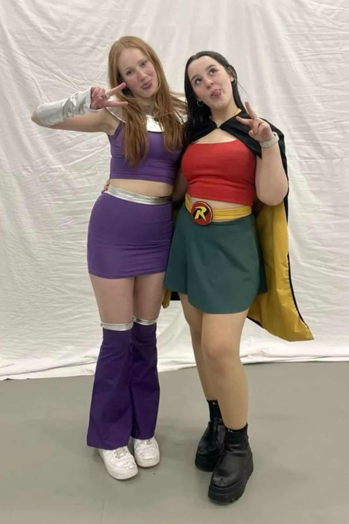 Two women in costumes pose in front of a white backdrop, both making peace signs. One wears a purple outfit with silver accents; the other wears a red, green, and yellow superhero costume.