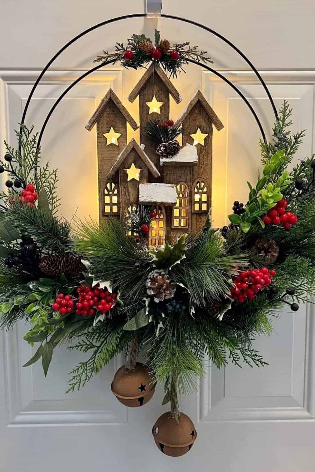 A holiday wreath features wooden houses with lit star windows, greenery, red berries, pinecones, and two hanging bells, displayed on a white door.