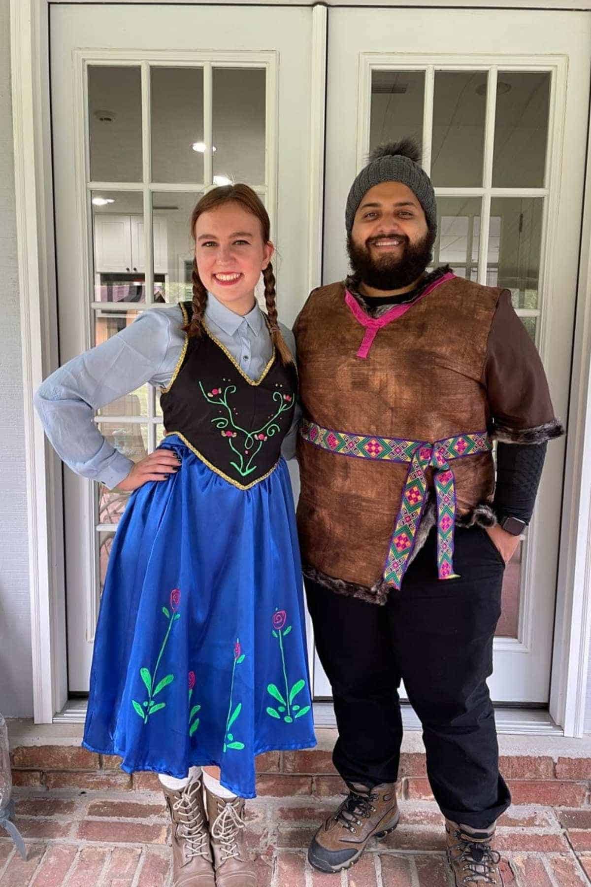 Two people stand in front of glass doors wearing costumes inspired by characters from the movie Frozen, with embroidered patterns and boots.