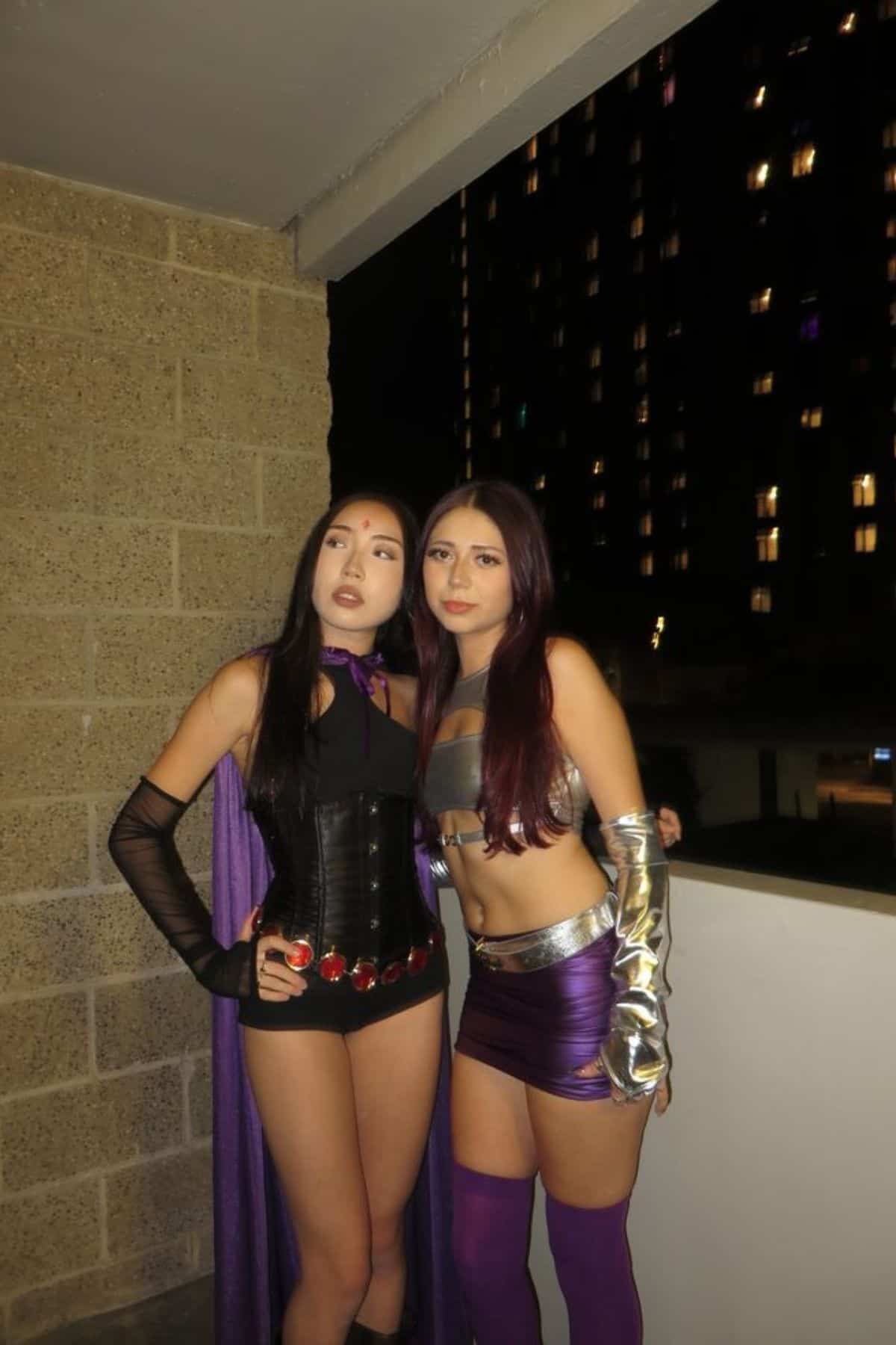 Two women in superhero costumes stand side by side on a balcony at night, with a city building lit up in the background.