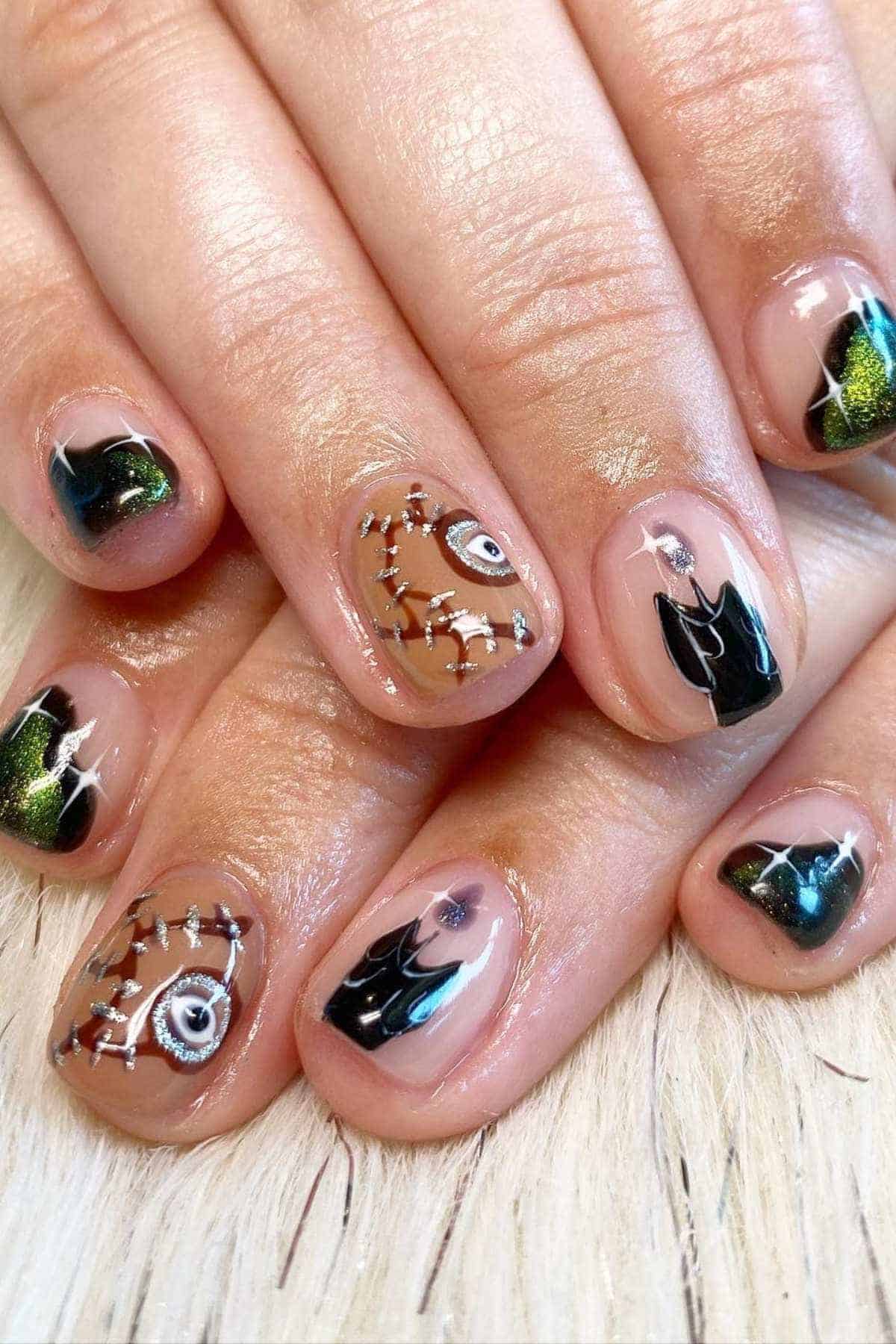Fingernails painted with a Halloween theme, featuring stitched patterns, eyes, black shapes, and shimmering green details on a natural base.