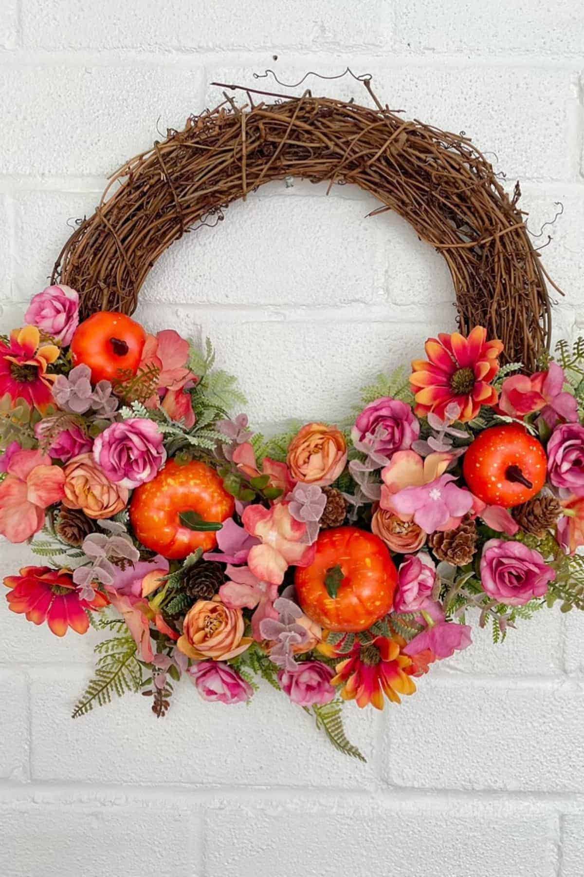 A grapevine wreath decorated with artificial pink flowers, greenery, and small orange pumpkins, hanging on a white brick wall.