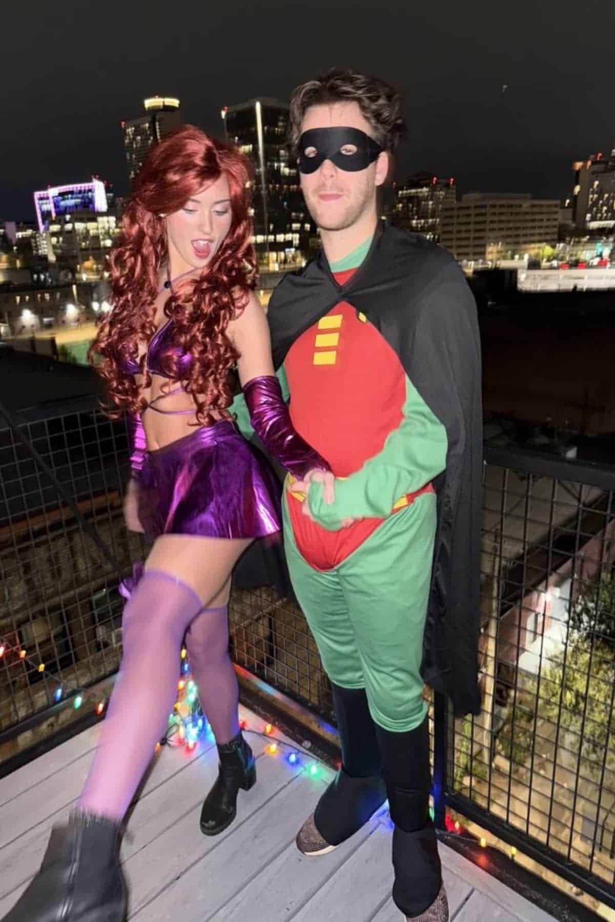 Two people in superhero costumes stand on a balcony at night with city lights in the background. One wears a purple outfit and wig; the other wears a black mask, cape, and green suit.