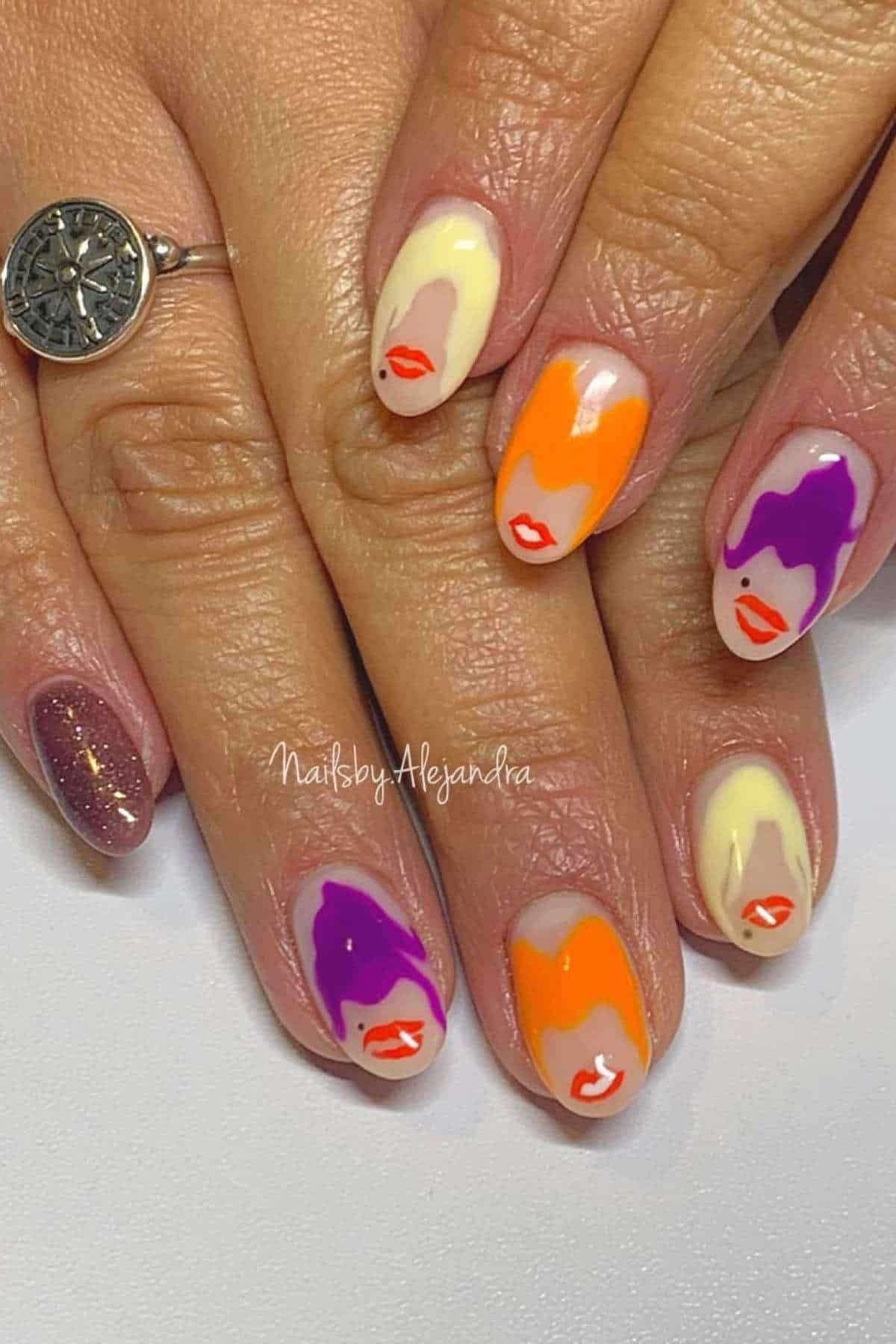 Hand with almond-shaped nails featuring abstract purple, orange, and cream designs with small red lips. One finger has a glittery purple polish. A silver ring is worn on one finger.