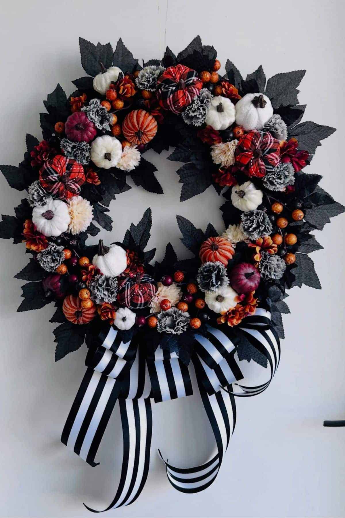 A wreath made of black leaves, small white and orange pumpkins, pine cones, berries, and a black and white striped ribbon bow.