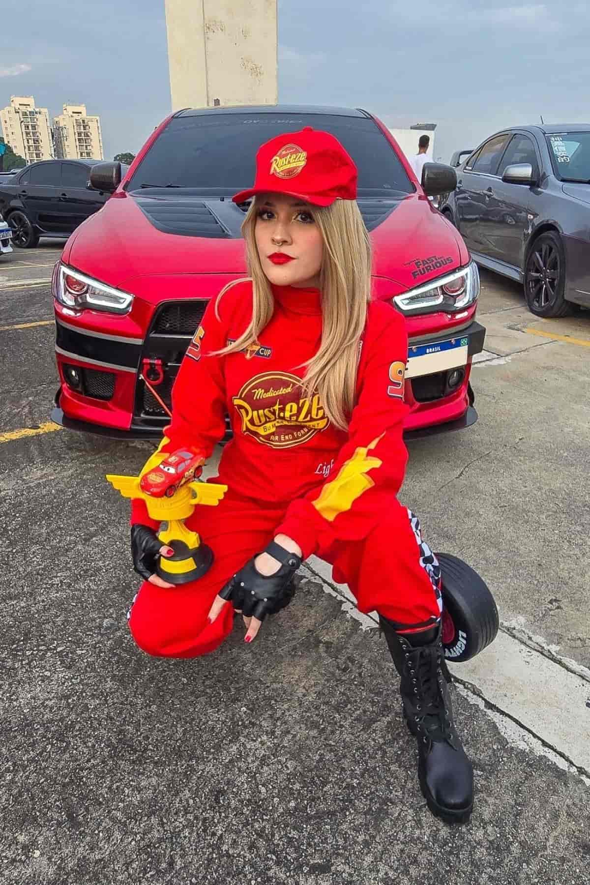 Person in a red racing costume and cap crouches in front of a red sports car in a parking lot, holding a yellow toy car.