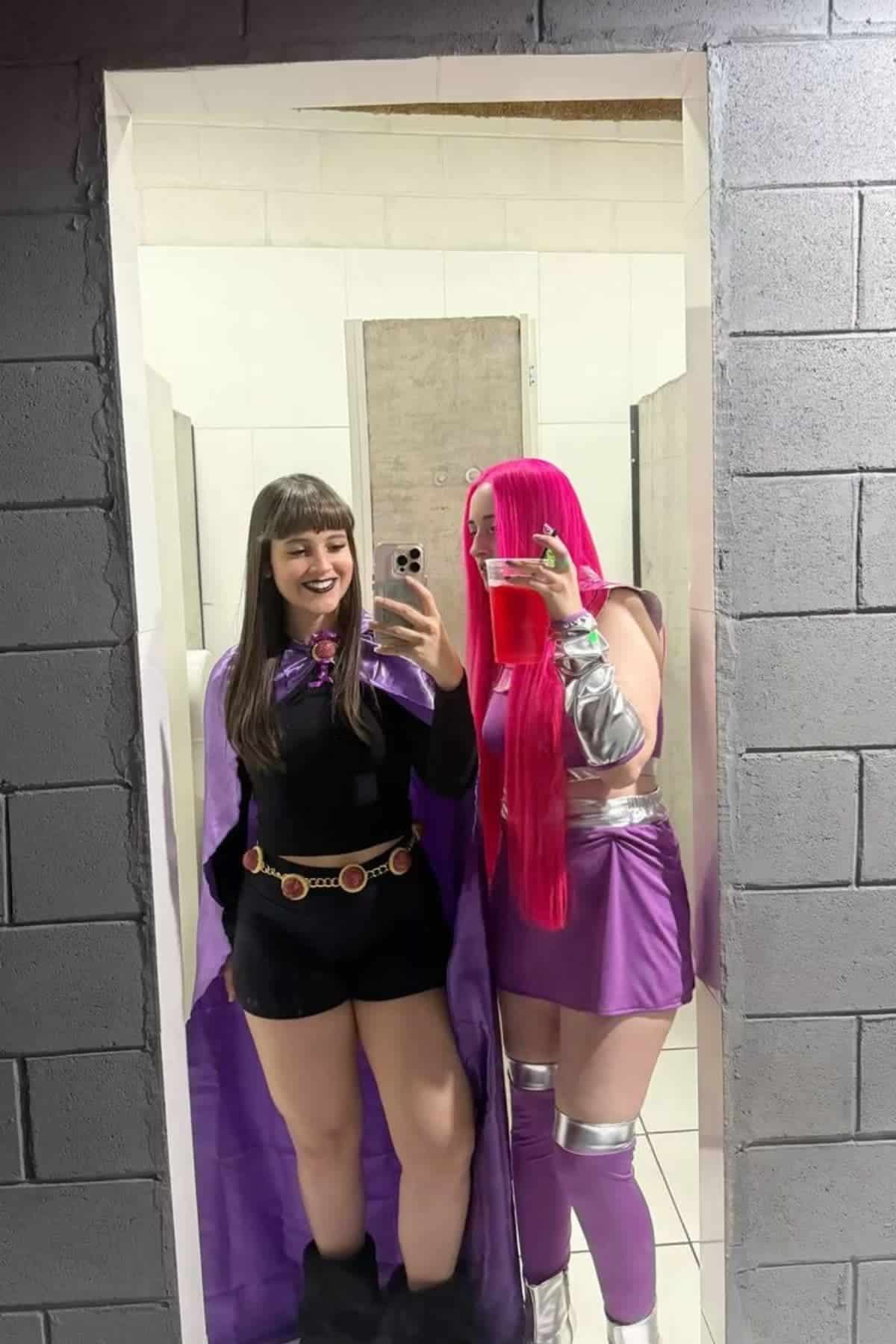 Two people in colorful superhero costumes take a mirror selfie in a bathroom; one has black hair and a cape, the other has long pink hair and holds a red cup.