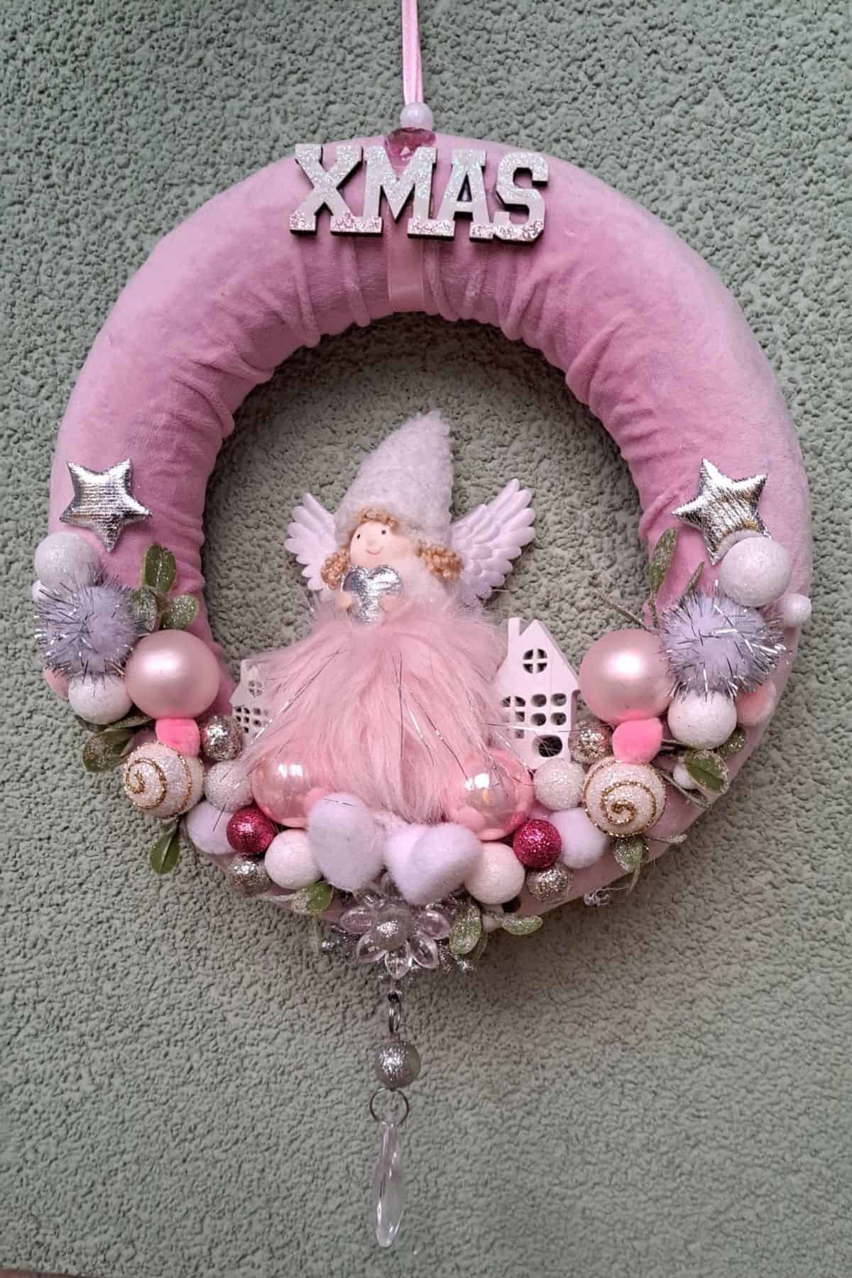 A pink Christmas wreath with “XMAS” letters, an angel figurine, small houses, baubles, stars, and decorative beads, hanging on a textured wall.