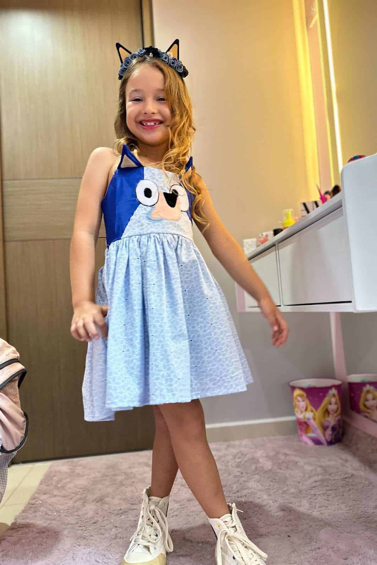 Young girl wearing a blue cartoon-themed dress and headband with cat ears, standing and smiling in a bedroom with pink decor and princess-themed bins.