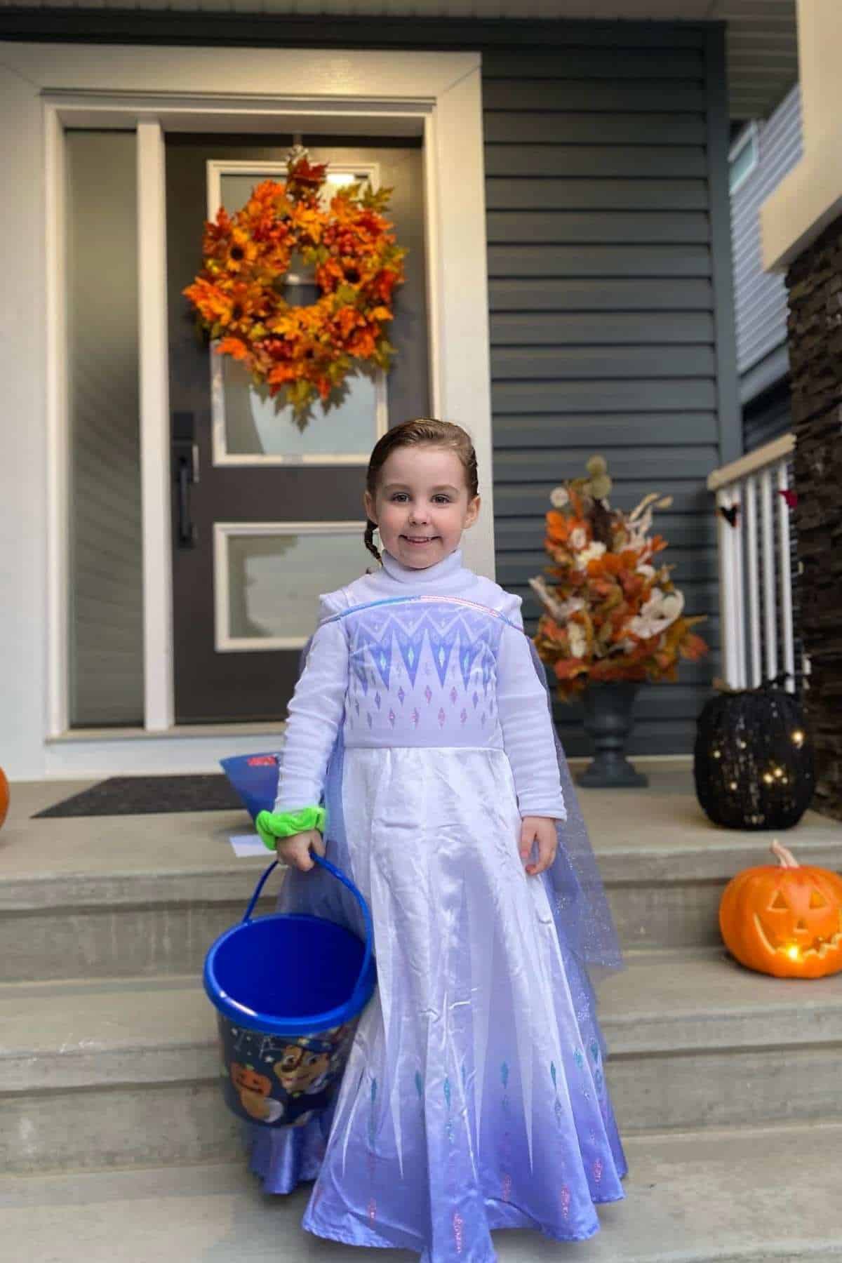A child in an ice queen costume stands on a porch holding a blue bucket, surrounded by pumpkins, autumn wreaths, and seasonal decorations.