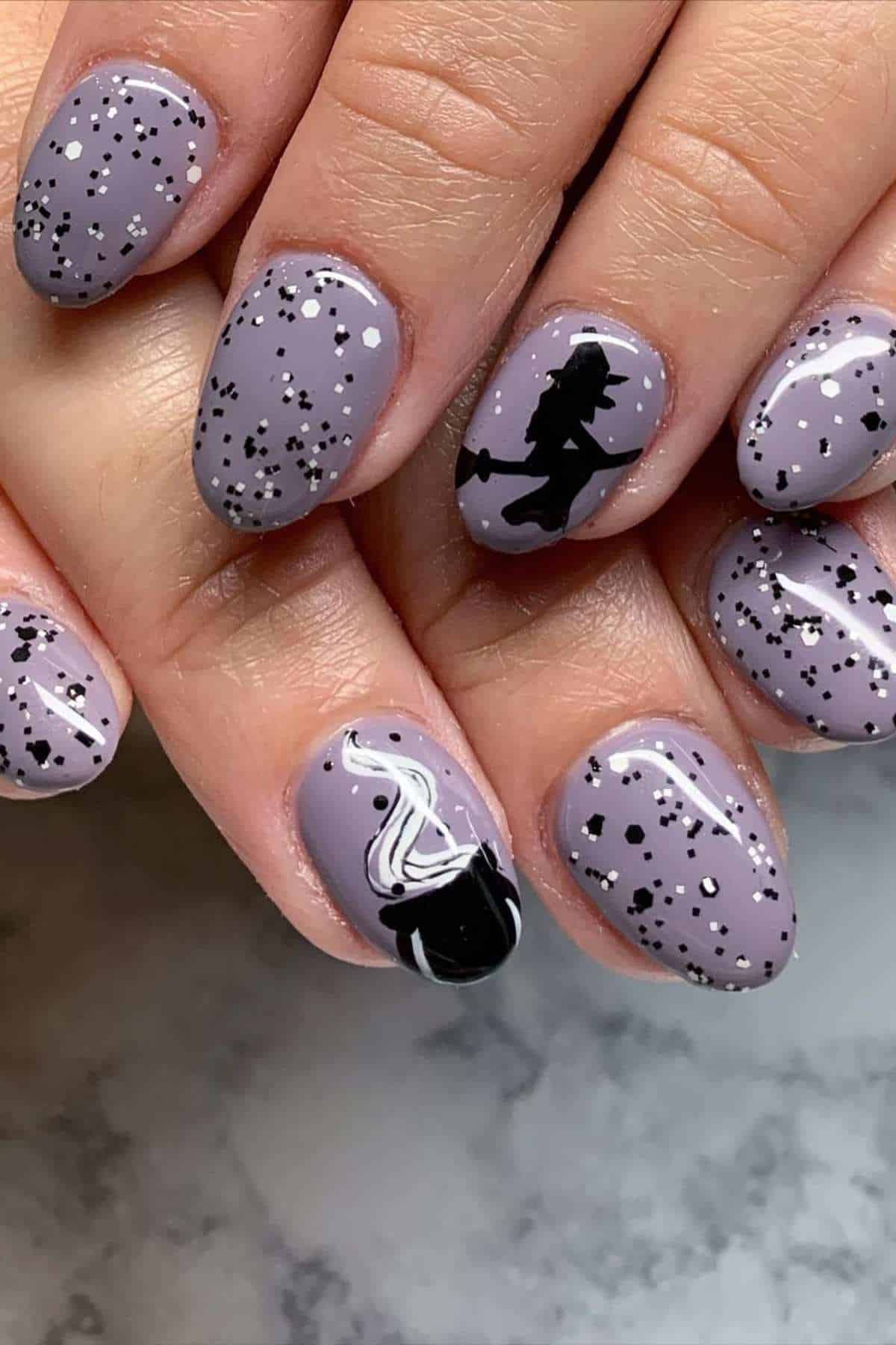 Close-up of hands with mauve nails featuring black and white speckles, a silhouette of a witch on a broomstick, and a nail art design of a black cauldron with white smoke.