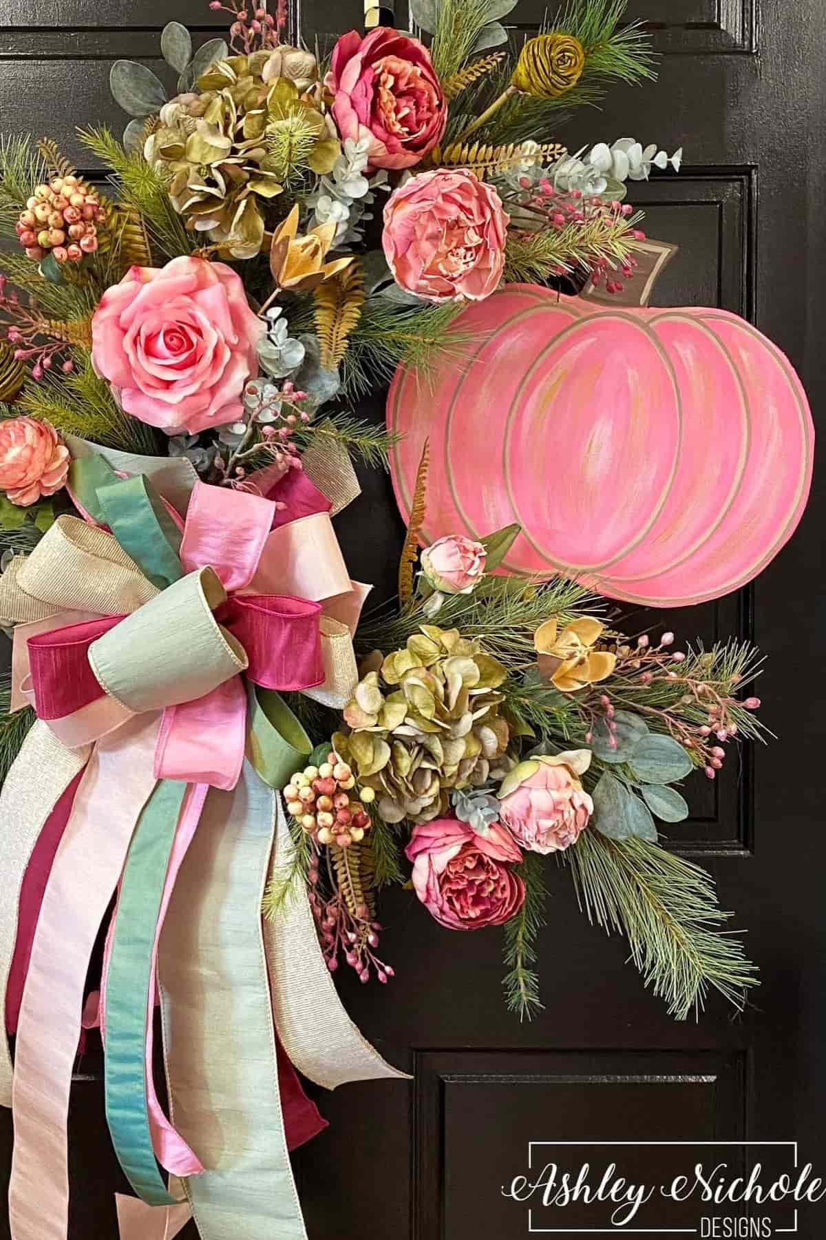 A floral wreath with pink and gold flowers, greenery, and pastel ribbons is displayed on a black door with a pink pumpkin decoration.