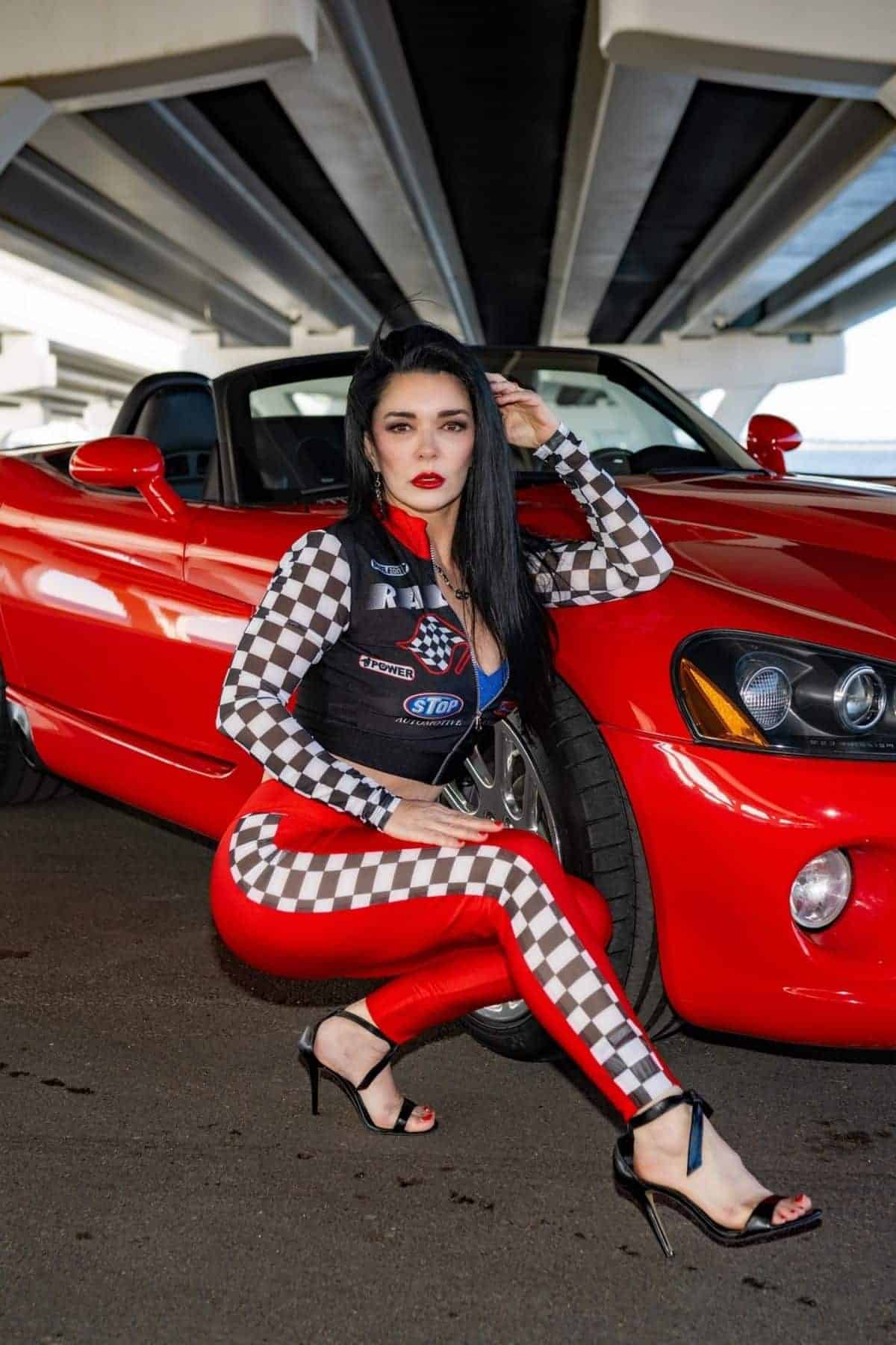 Woman in racing-themed outfit poses next to a red convertible under a bridge, resting her hand on the car’s front wheel.
