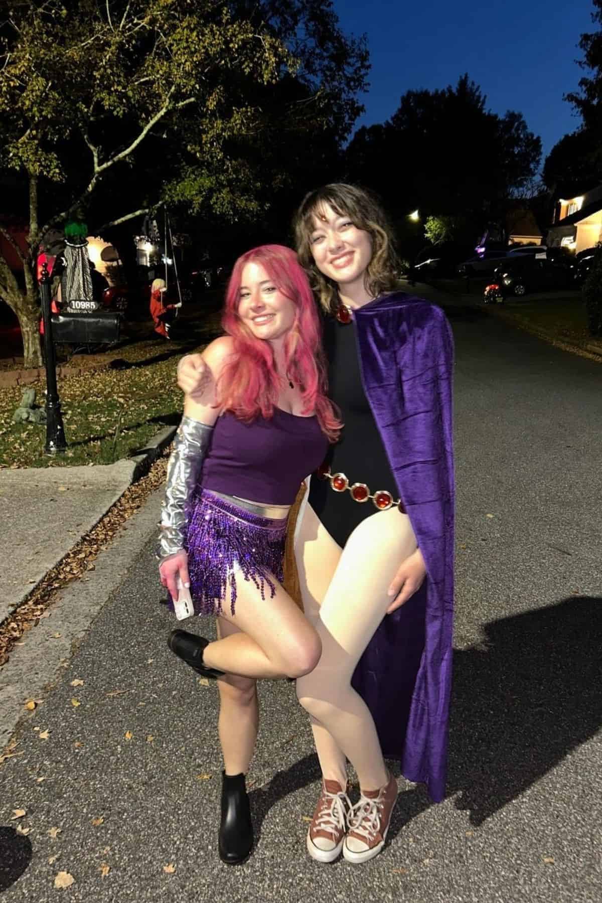 Two people in costume stand outdoors at night, smiling and posing together on a street lined with trees and houses.