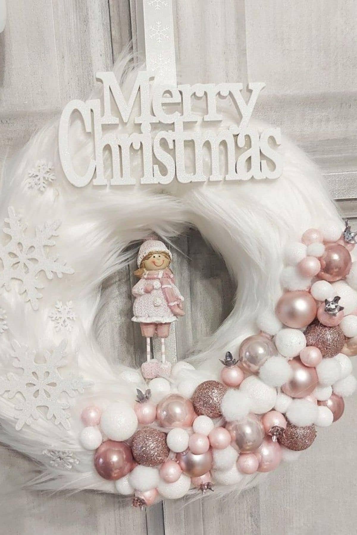 A white Christmas wreath with snowflake decorations, pink and white ornaments, a small doll, and a “Merry Christmas” sign on top, hanging on a light-colored wooden door.