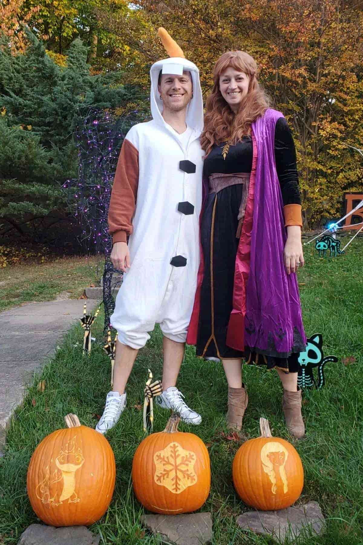 Two adults in Frozen-themed costumes stand outdoors behind three decorated pumpkins, with trees and Halloween decorations in the background.