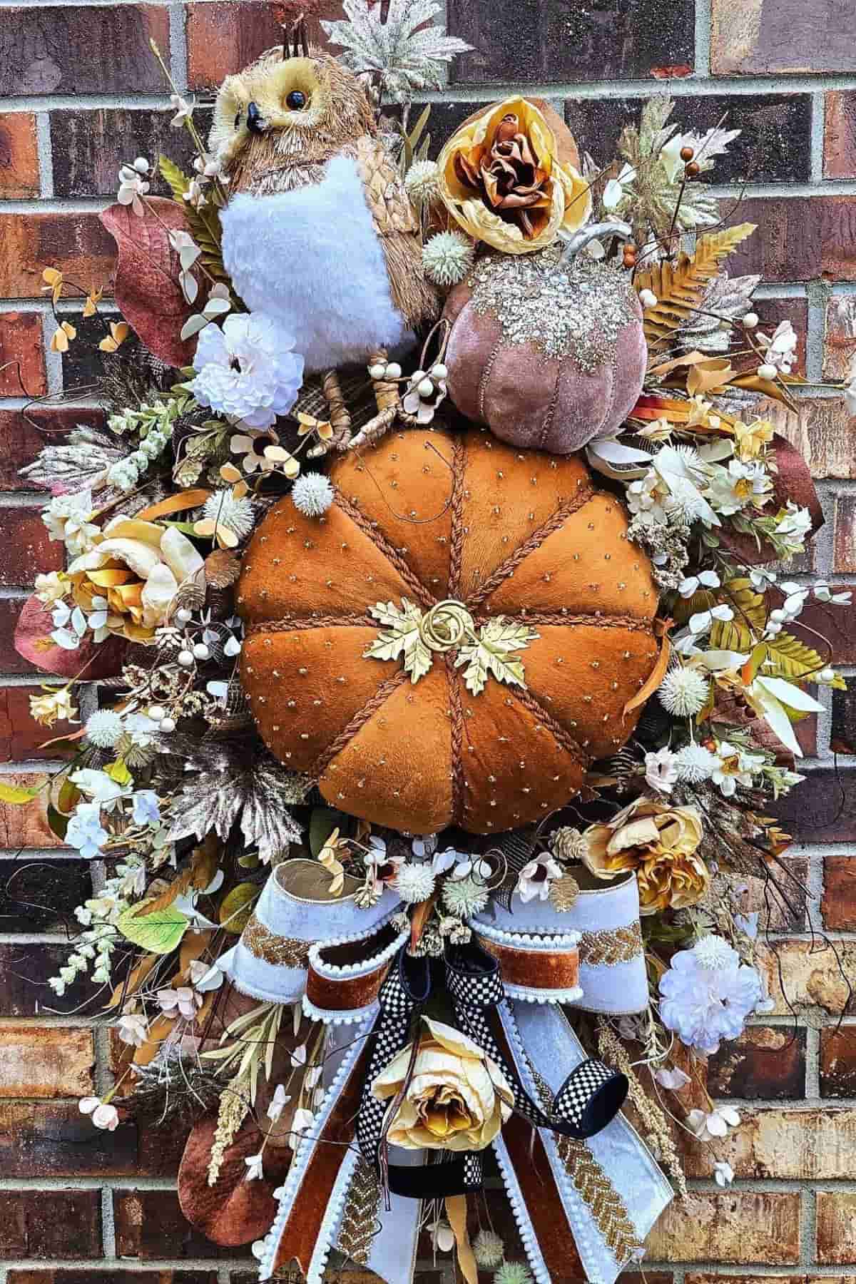 Autumn wreath with a plush owl, velvet pumpkin, artificial flowers, leaves, and a decorative bow, displayed against a brick wall.