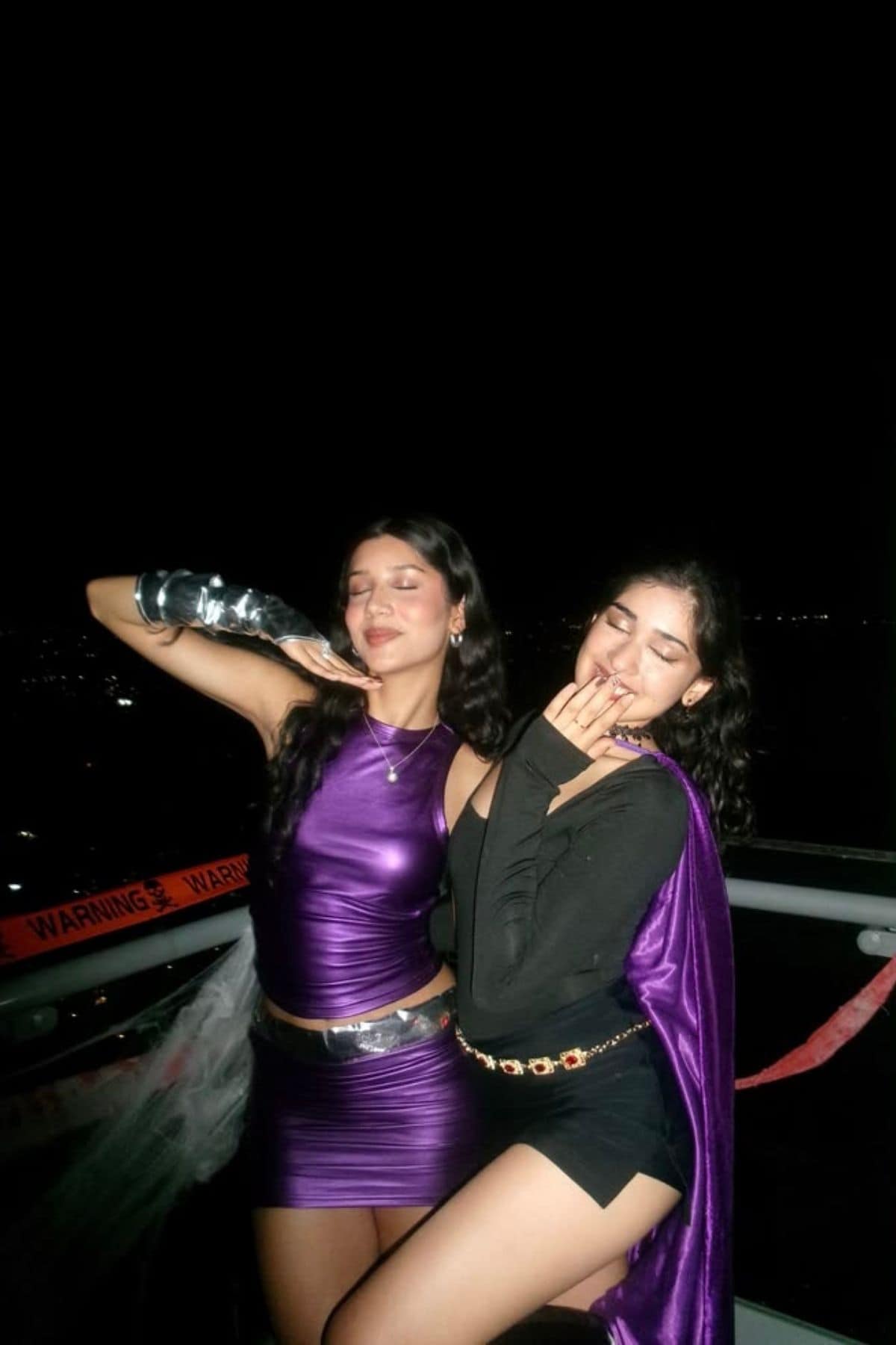 Two women in costumes pose on a balcony at night; one wears a shiny purple outfit with metallic armwear, the other wears black with a purple cape. Warning tape is visible in the background.