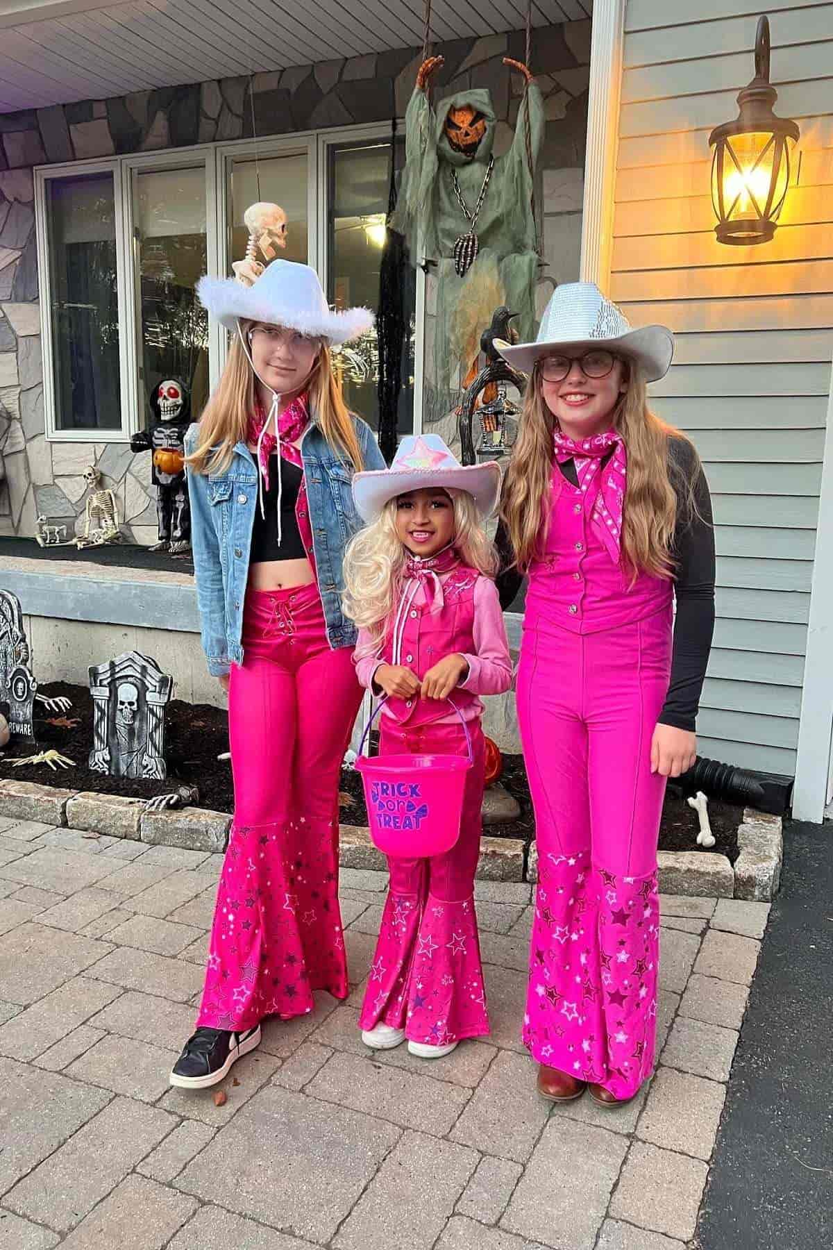 Three children in matching bright pink cowgirl costumes and hats stand outside a house decorated for Halloween with spooky props.