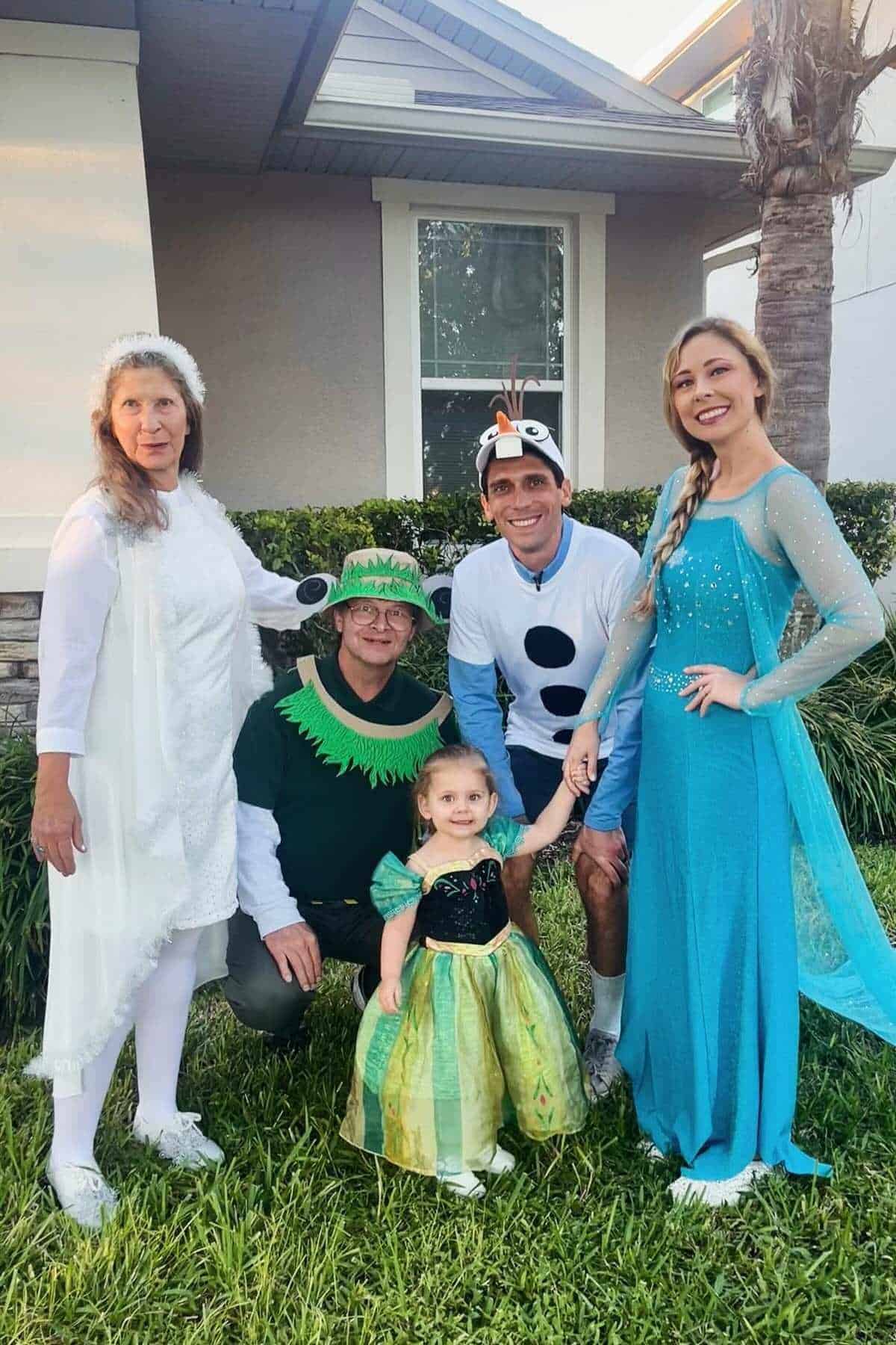 Five people pose outdoors in costumes, including a frog, a snowman, Elsa, Anna, and an angel, in front of a house with a window and a palm tree.