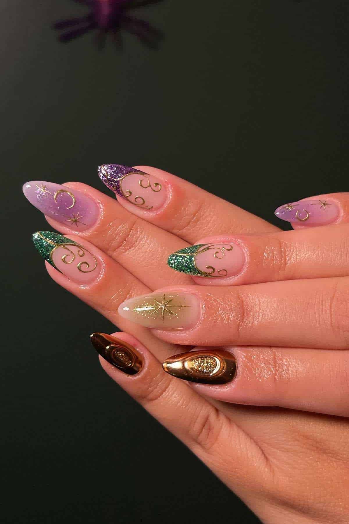 A hand with long, almond-shaped nails featuring purple, green, and gold designs with swirls, stars, and metallic accents against a dark background.