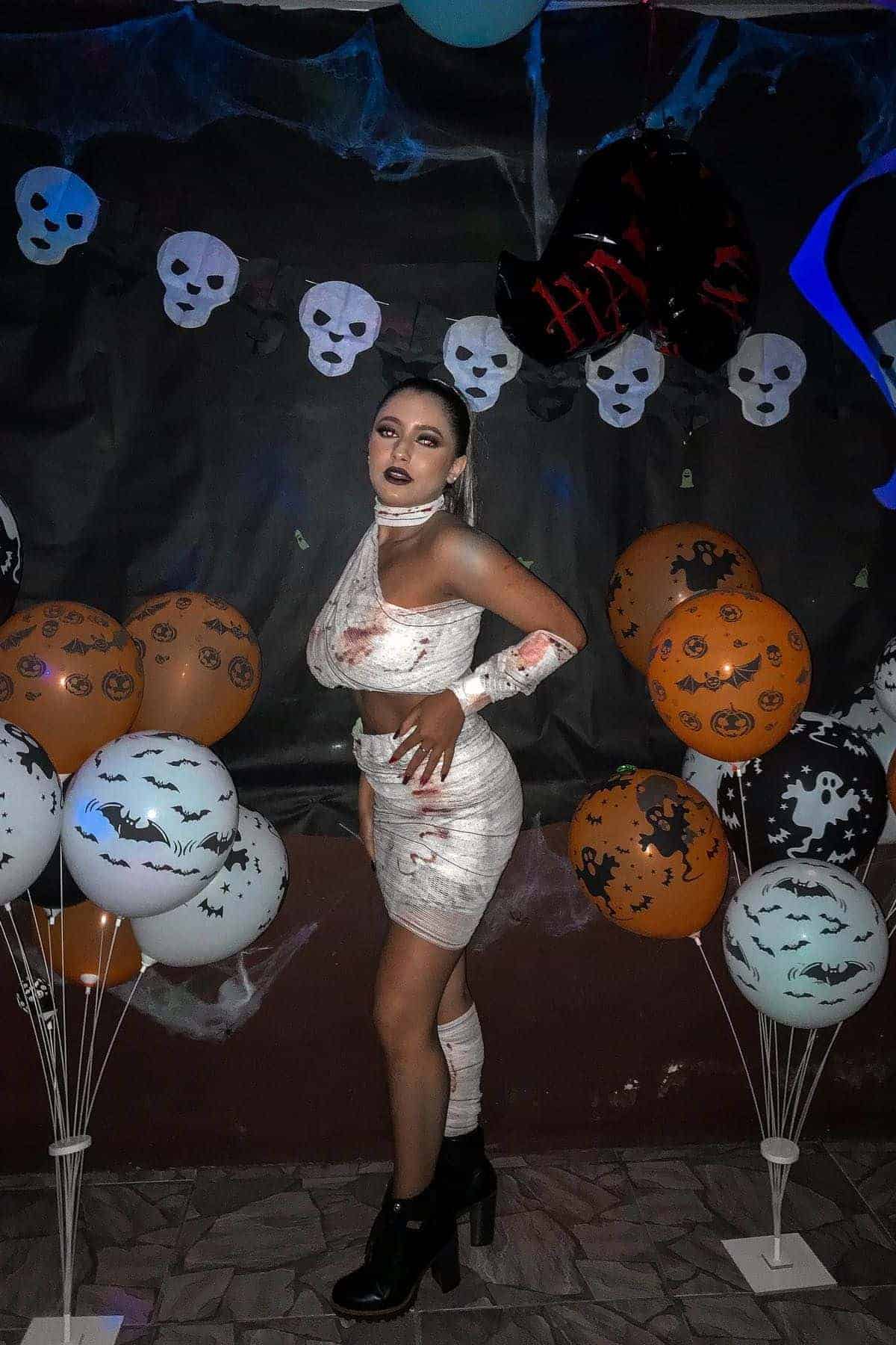 A person in a mummy costume poses at a Halloween party, surrounded by pumpkin and bat balloons, with skull decorations on the wall behind.