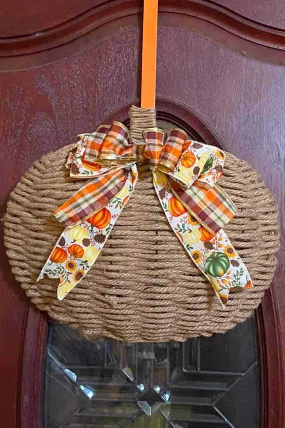A rope-wrapped pumpkin-shaped door decoration with plaid and pumpkin-patterned ribbon bows, hanging on a wooden door by an orange ribbon.