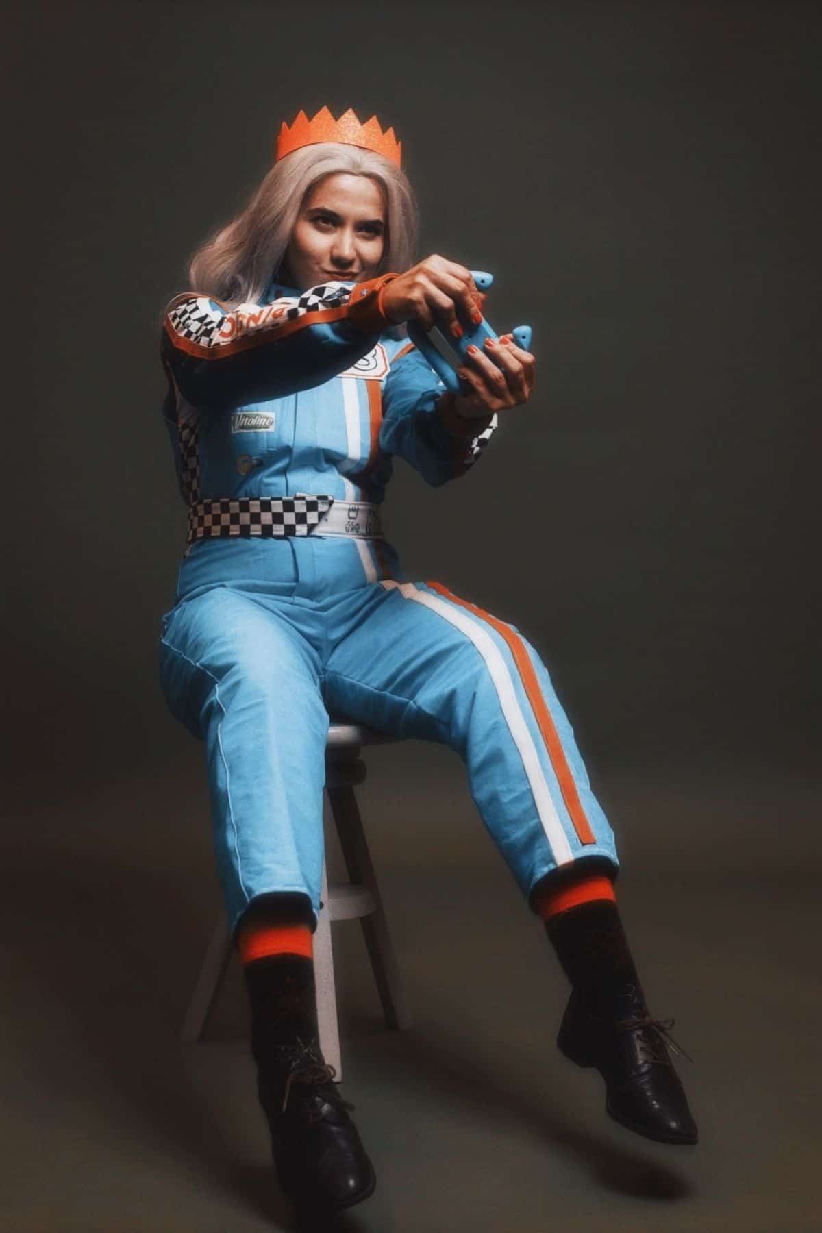 Person in blue racing jumpsuit and orange paper crown sits on a stool, holding a toy car, against a dark background.