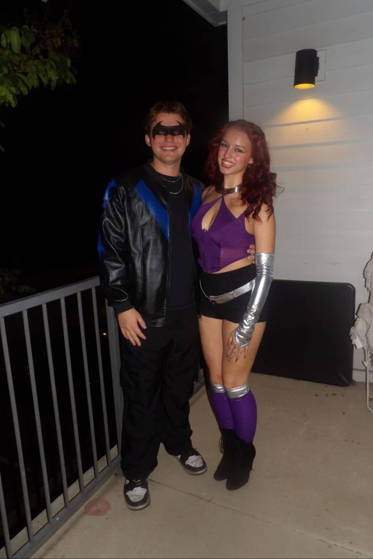 Two people in superhero costumes stand on a balcony at night, smiling at the camera; one wears a black outfit with a mask, the other a purple top, shorts, and silver arm bands.