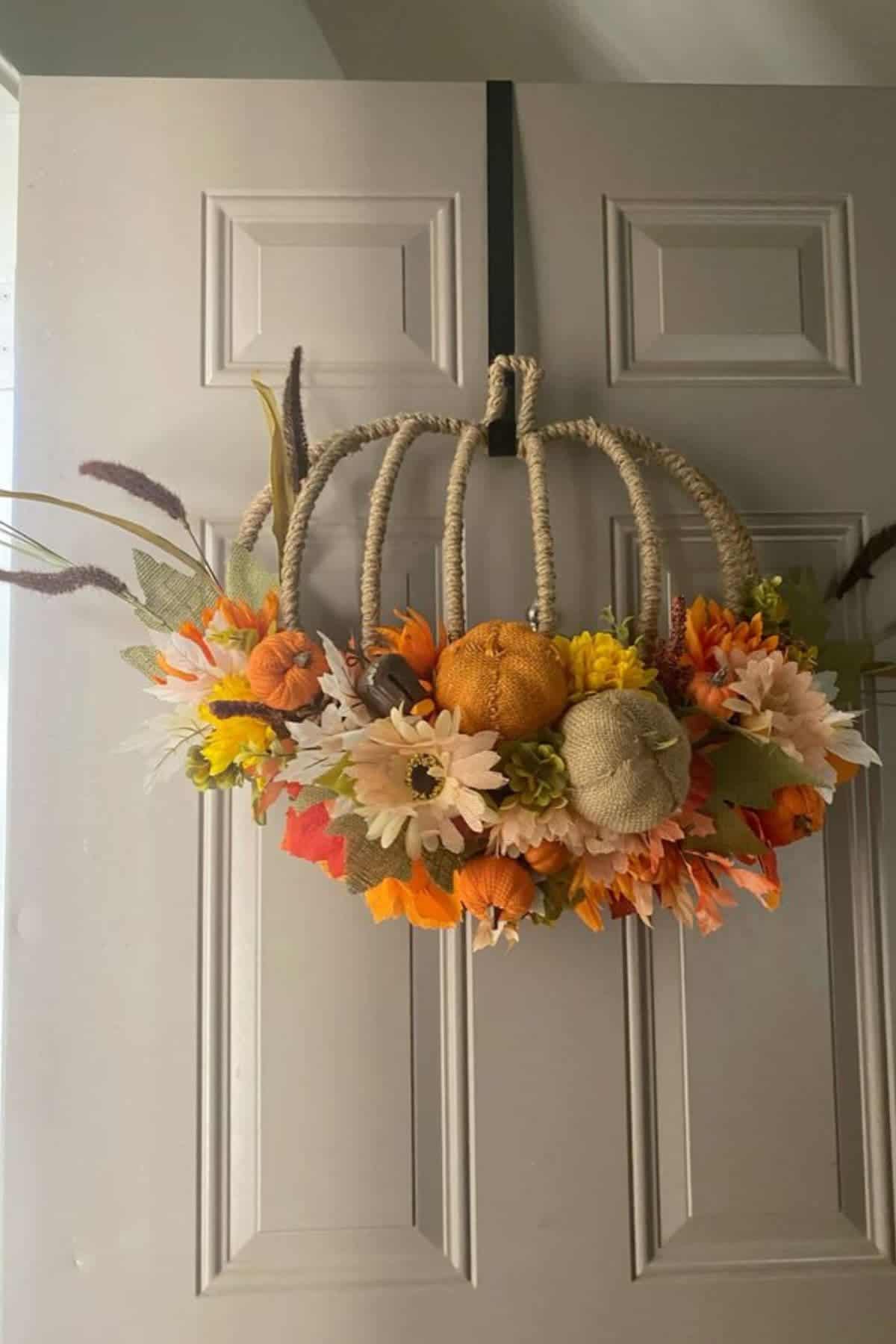 Wreath shaped like a pumpkin, decorated with artificial flowers and mini pumpkins in autumn colors, hangs on a beige door.