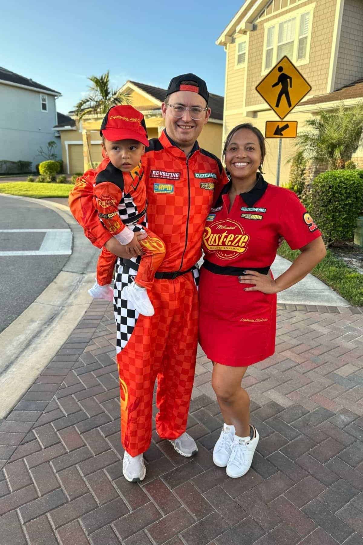 Three people in matching red racing-themed outfits stand on a suburban street, with houses and a pedestrian crossing sign in the background.